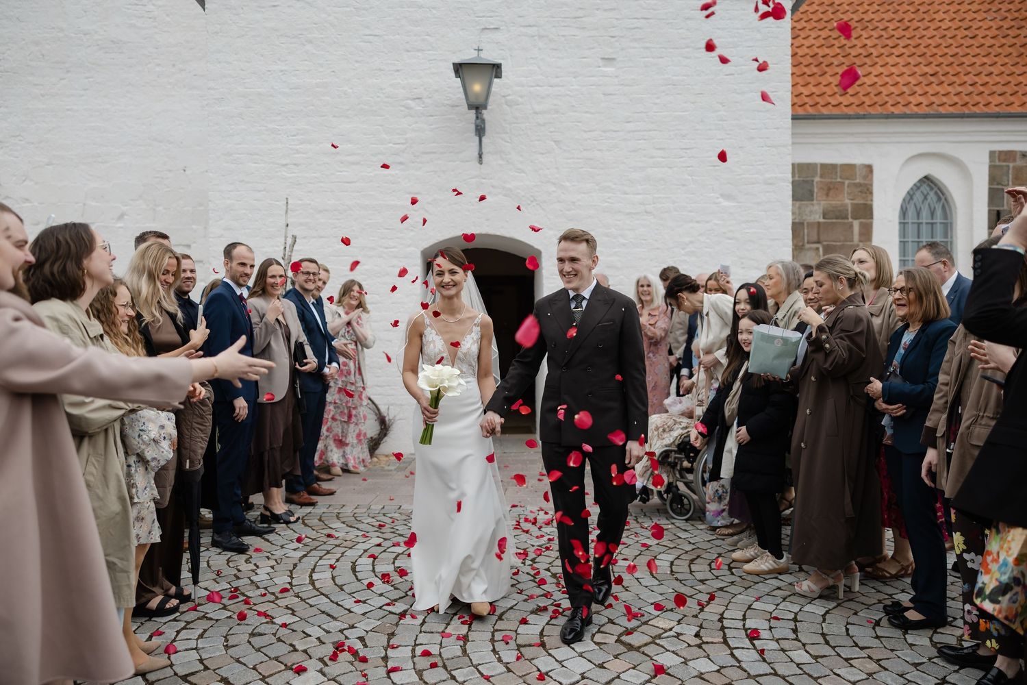 Bride and groom walk through rose petal confetti outside a white church surrounded by guests.