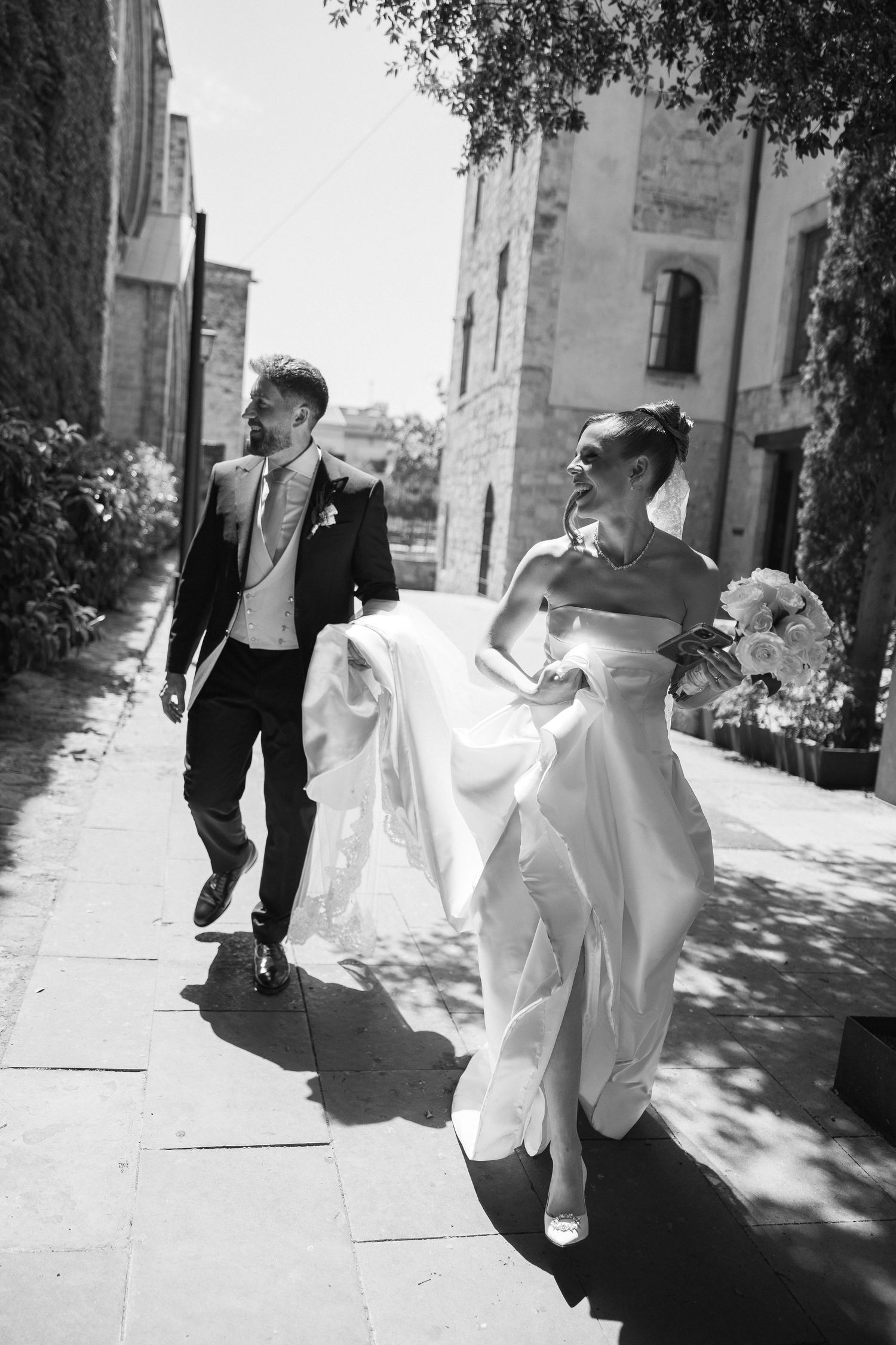 Black and white photo of a bride and groom walking together on a sunny cobblestone path beside historic stone buildings.