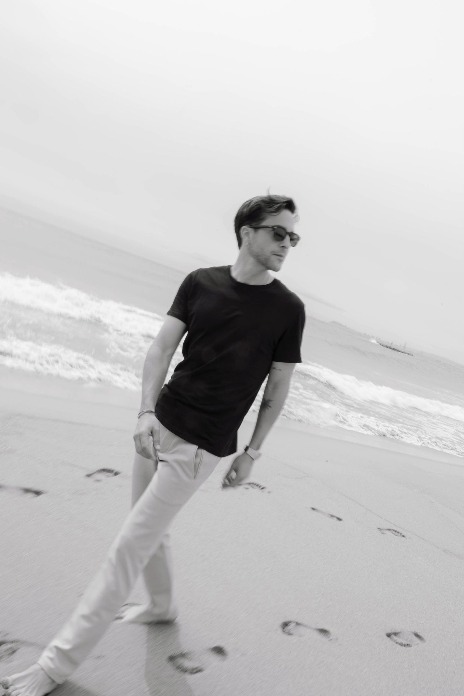 Man in black t-shirt and white pants strolling along a sandy beach wearing sunglasses in black and white.