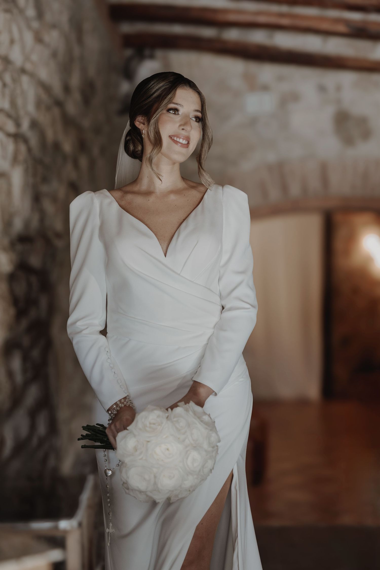 Smiling bride in elegant long-sleeve white wrap wedding dress holding peony bouquet in rustic venue.