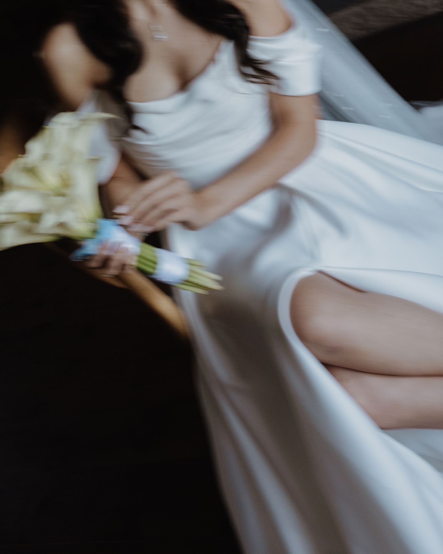 Blurred artistic photo of bride in white off-shoulder gown holding white calla lily bouquet.