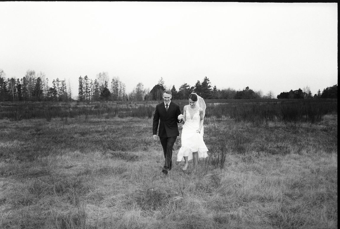 Black and white film photo of a bride and groom walking through an open field on their wedding day.