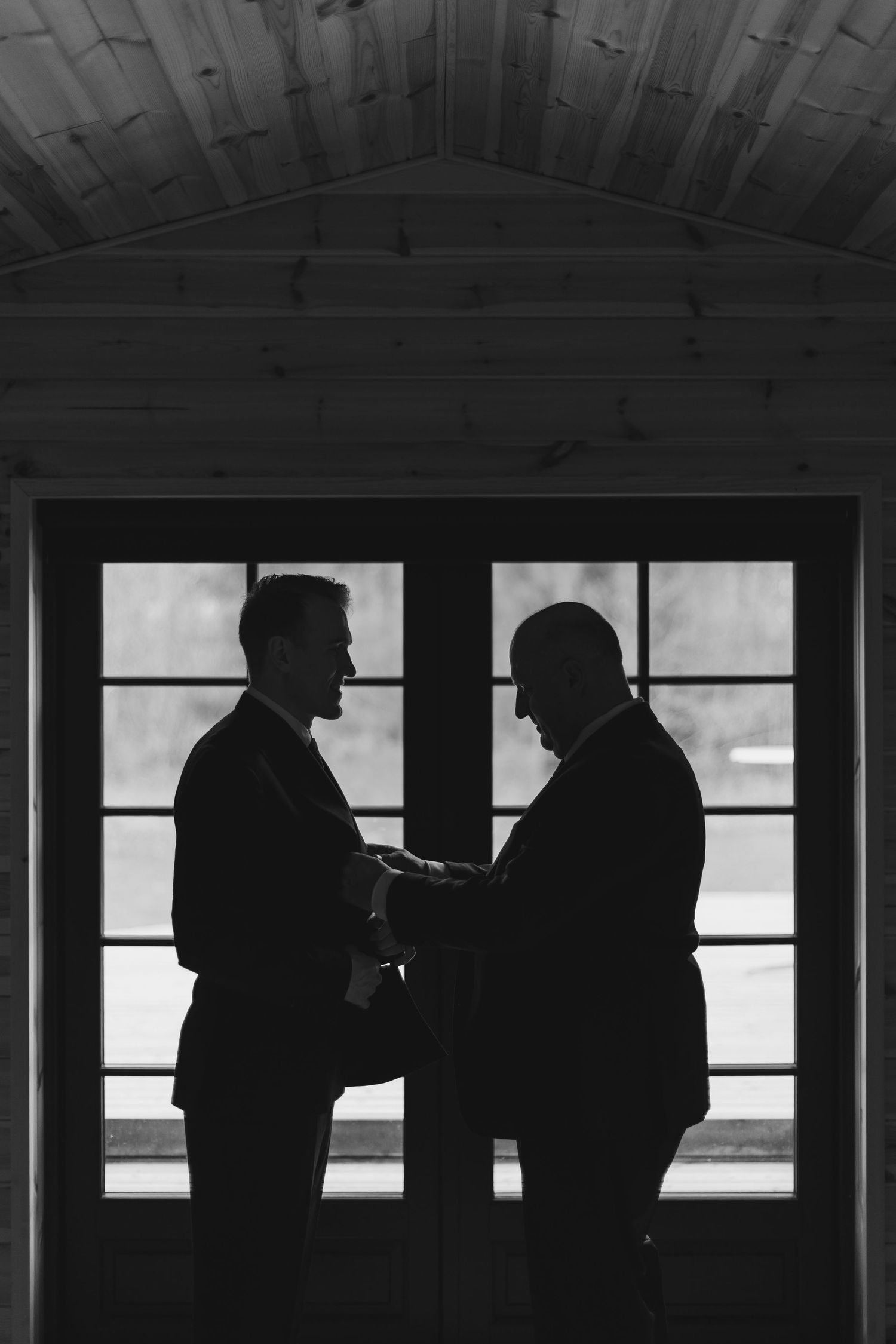 Two men in suits share an intimate moment as silhouettes against French doors in a black and white wedding photo.