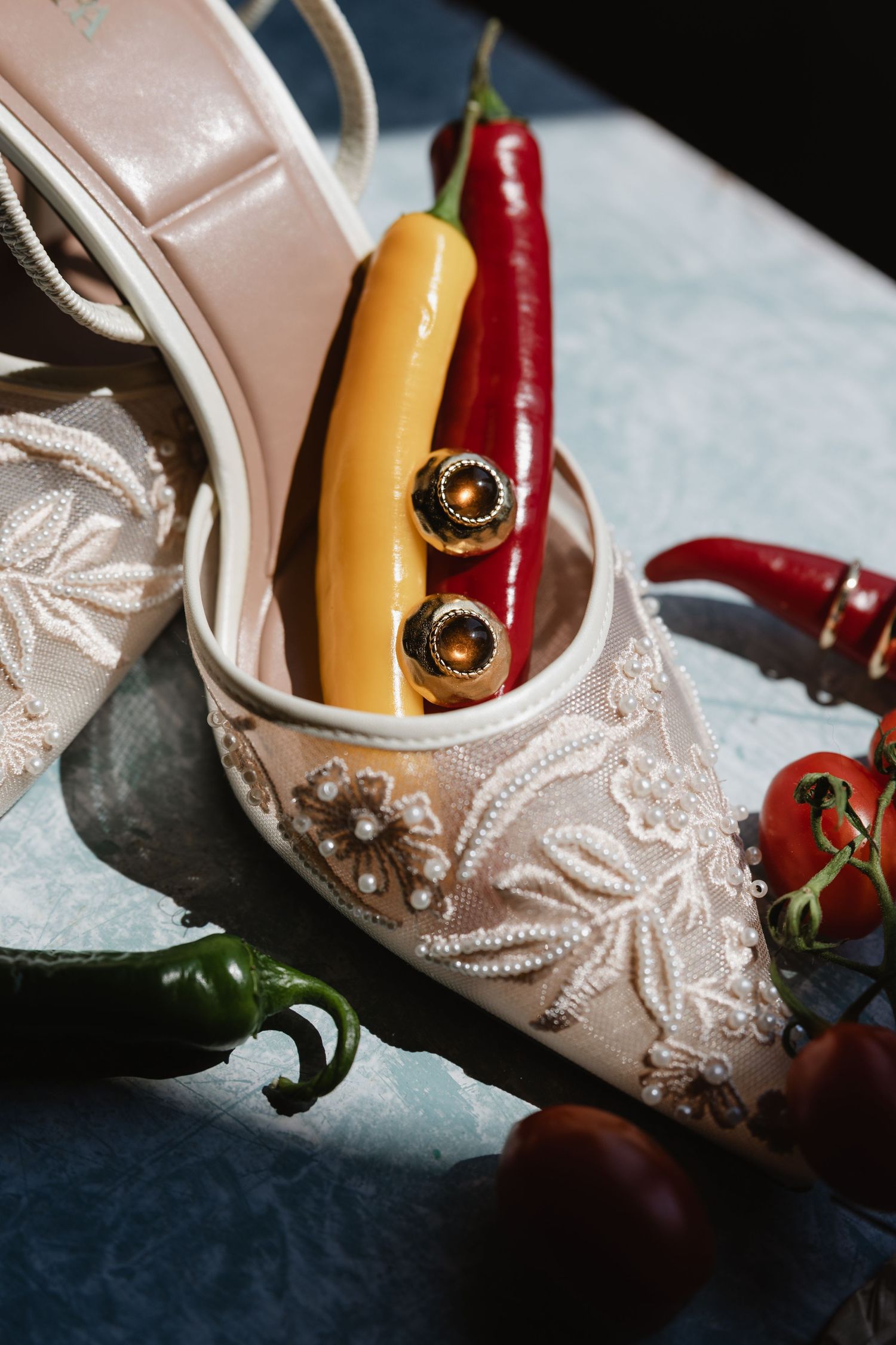 Elegant white lace bridal heels with pearl embroidery displayed with gold earrings, red, yellow, green chili peppers and tomatoes.