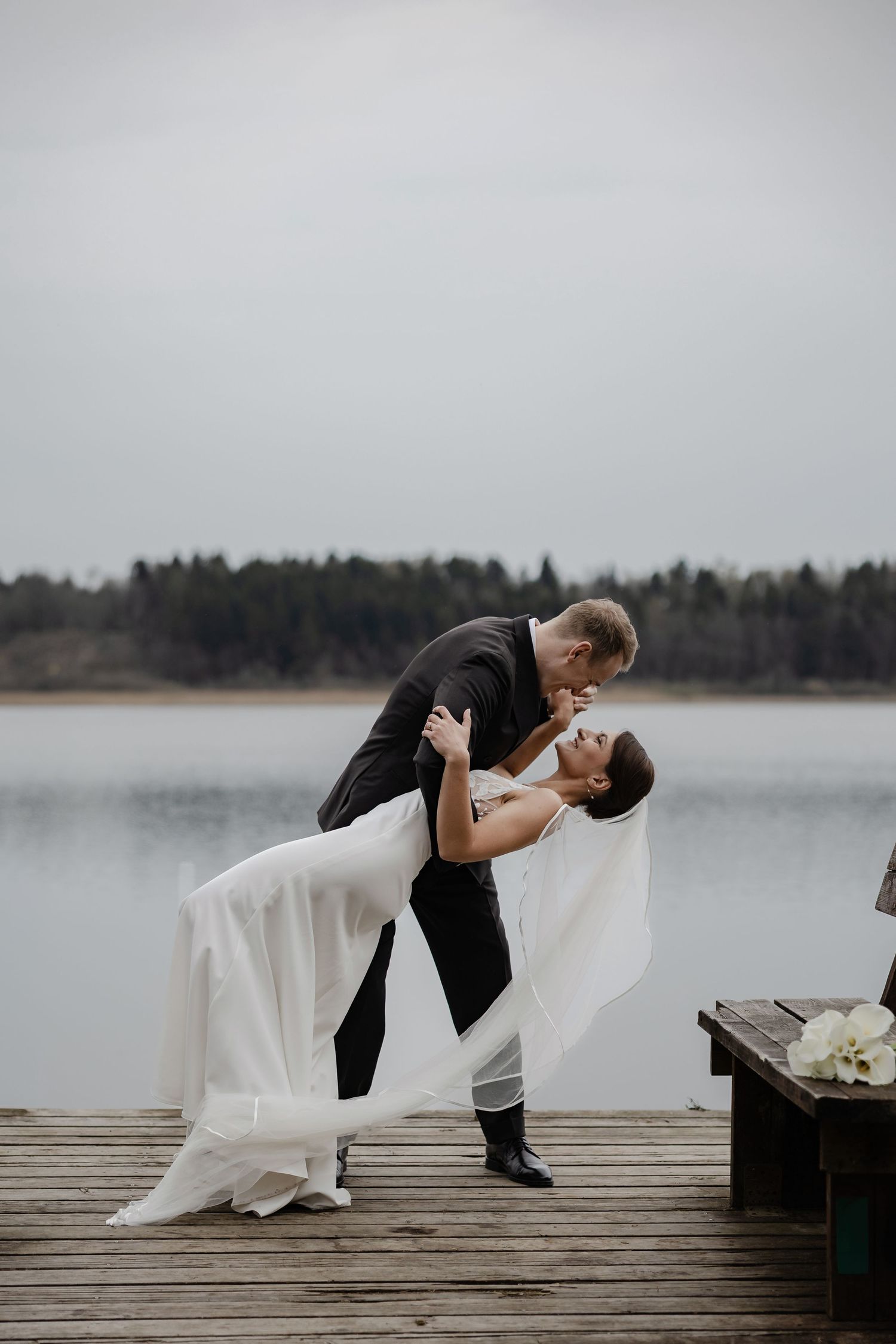 Groom dips bride in a romantic kiss on a wooden dock overlooking a calm lake on their wedding day.