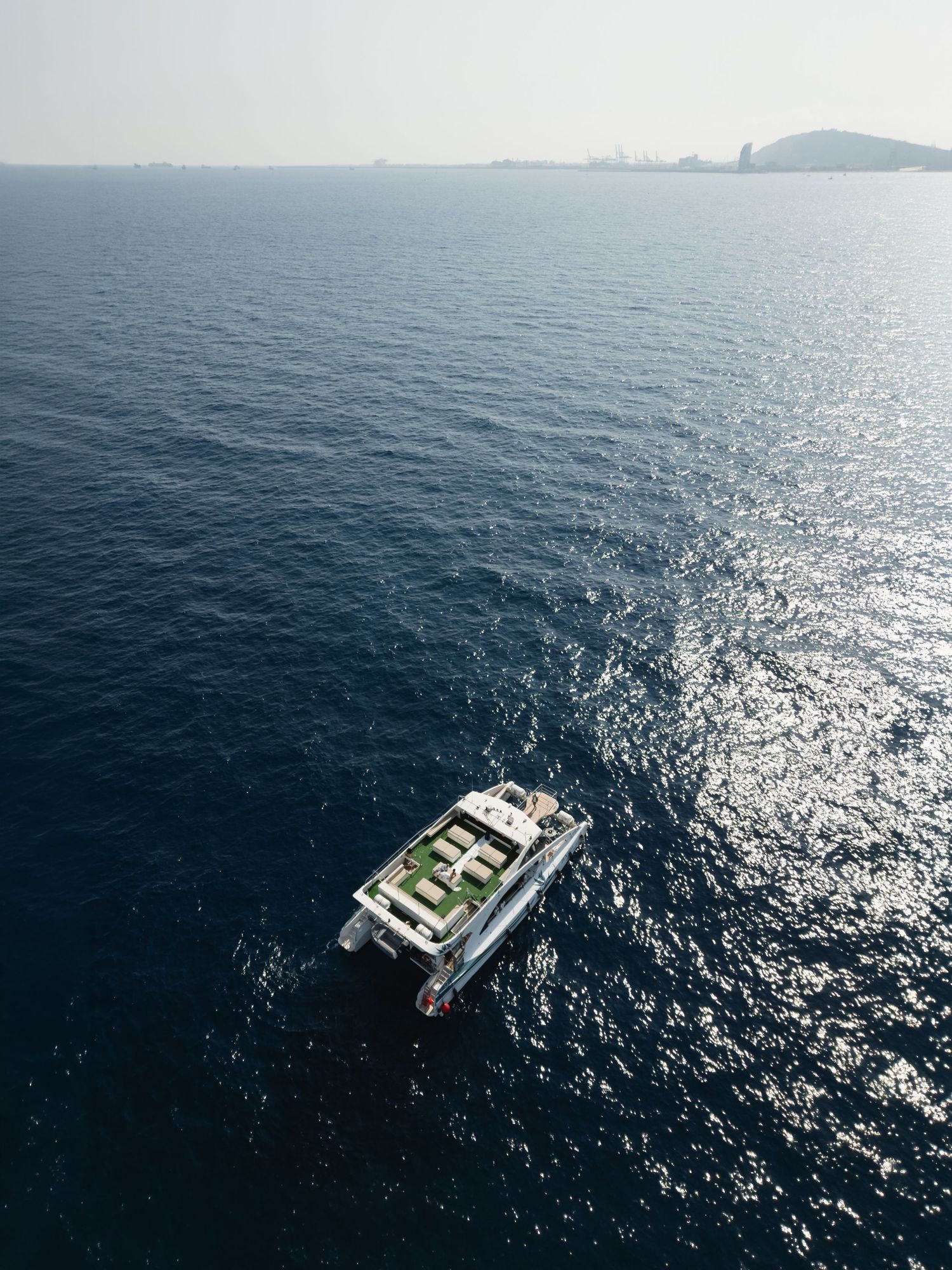 Aerial view of a luxury white yacht sailing on calm deep blue ocean waters with a hazy coastline in the background.