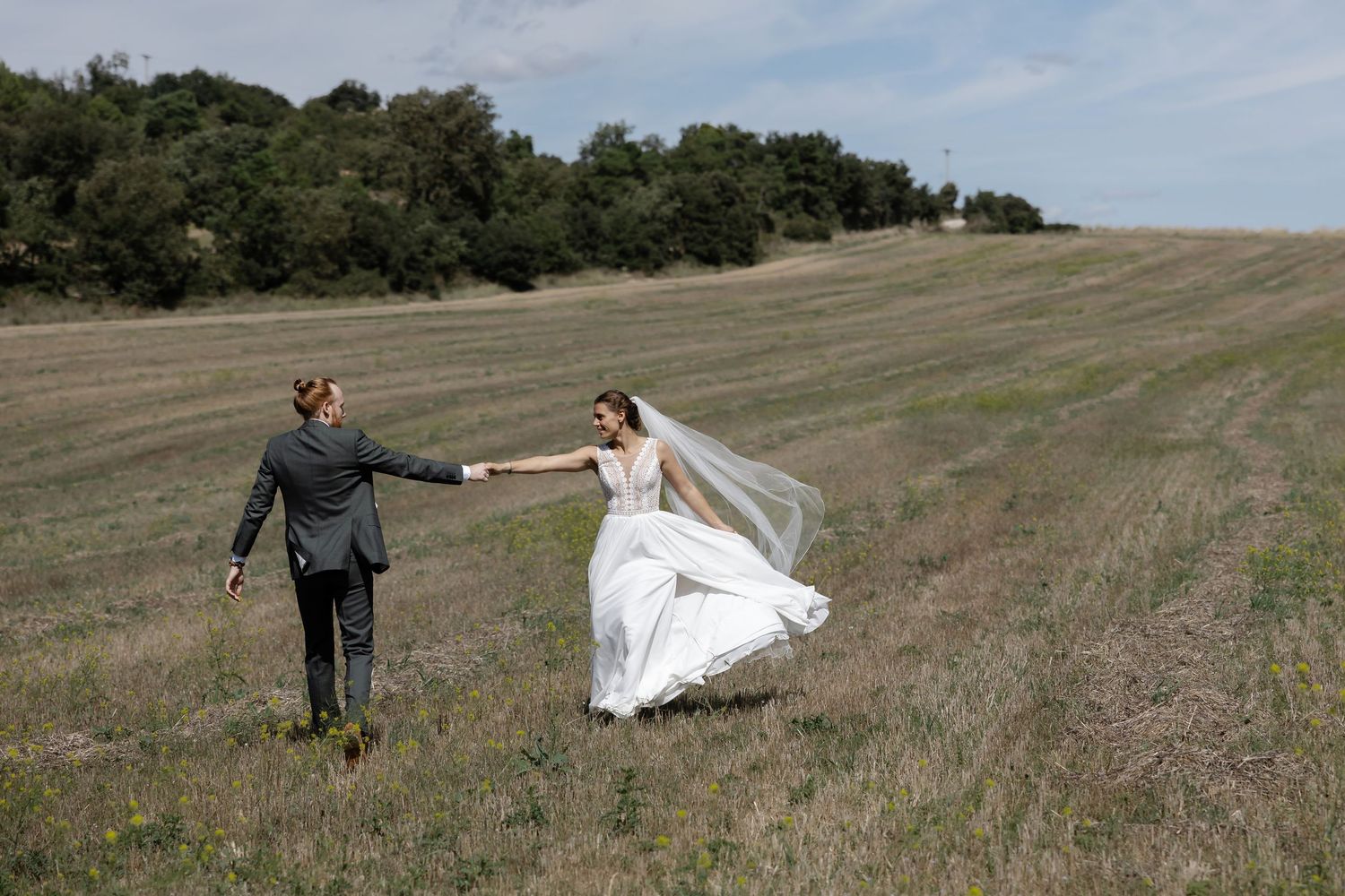 Bride and groom holding hands walking through a golden grass field, wedding veil flowing in the wind.
