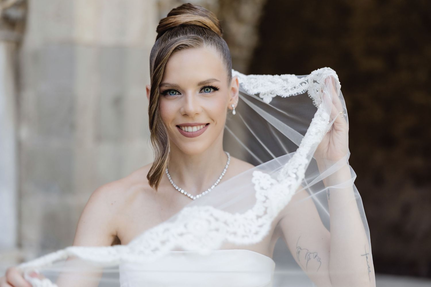 Smiling bride in white dress holding lace veil, wearing pearl necklace and earrings on her wedding day.