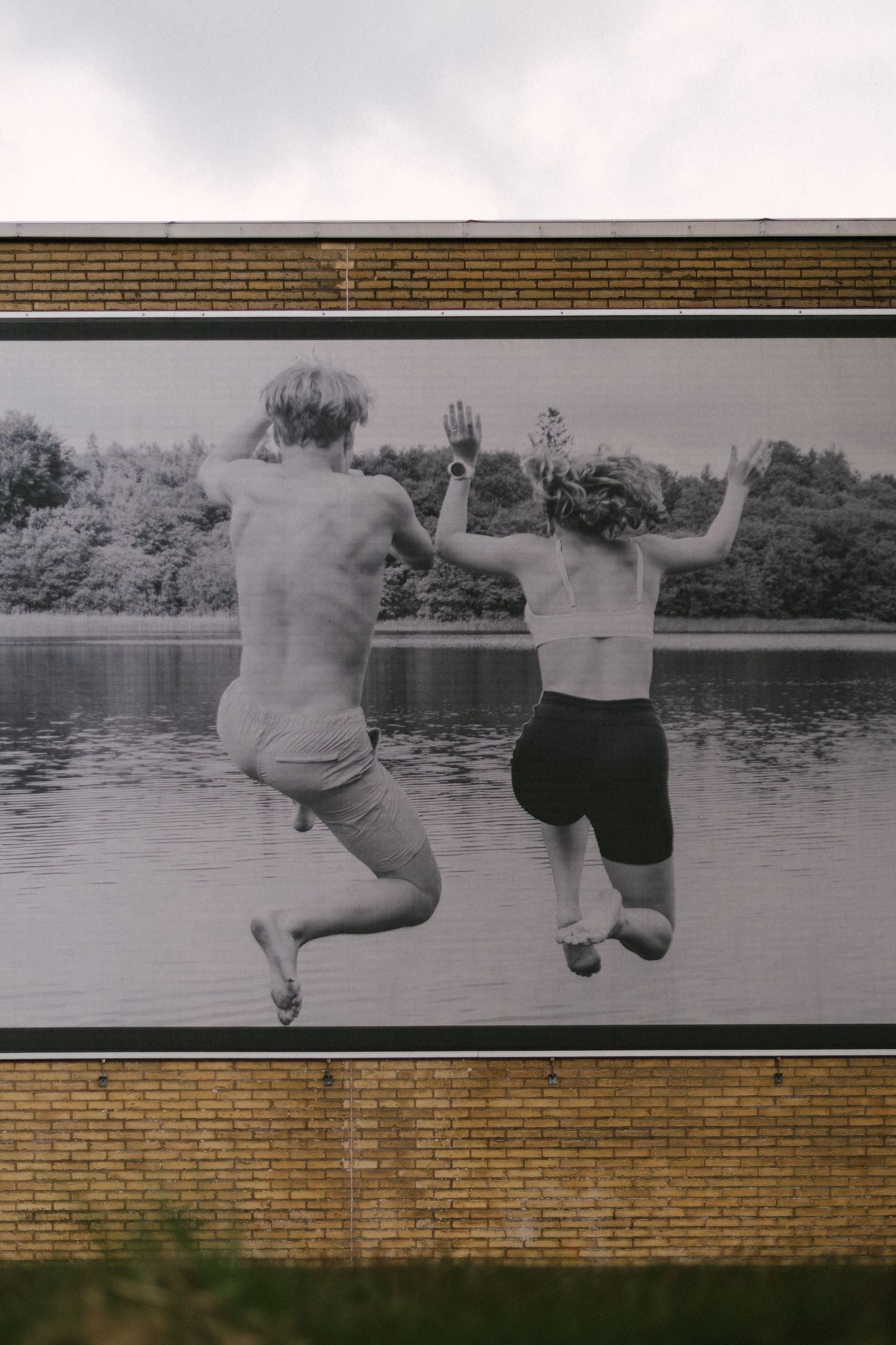 Black and white photo billboard of two people jumping into a lake, mounted on a brick wall outdoors.