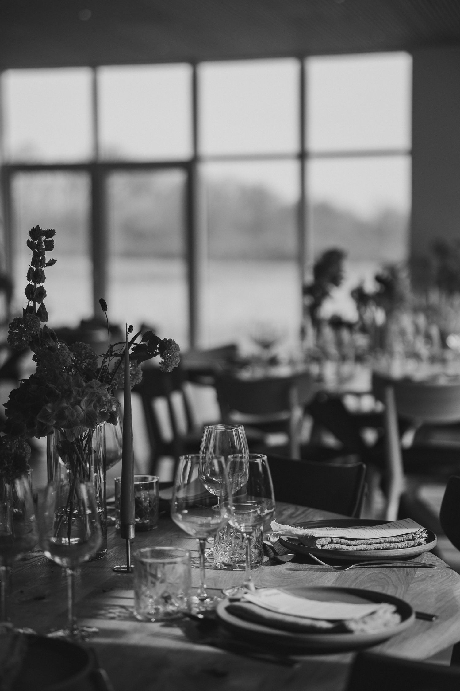 Elegant black and white wedding reception table setting with floral arrangements, wine glasses, and plates near large windows.