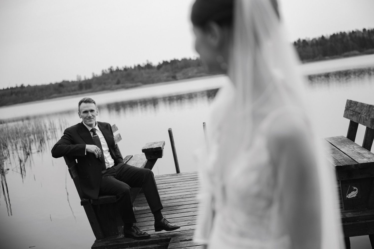 Black and white wedding photo of groom sitting on dock by lake, bride in veil standing in foreground.
