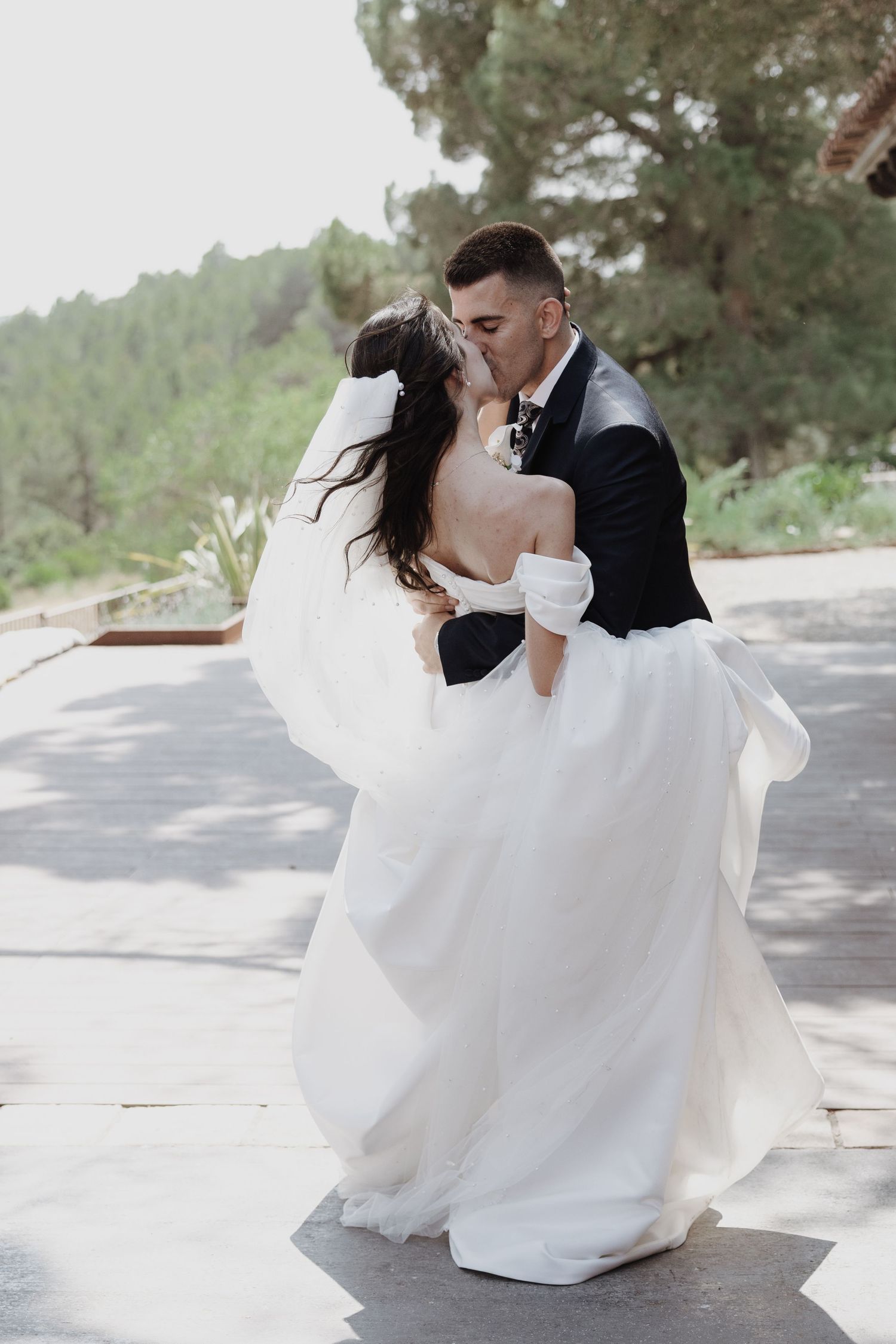 Bride and groom sharing a romantic kiss outdoors, with the bride in an elegant white off-shoulder gown surrounded by lush greenery.