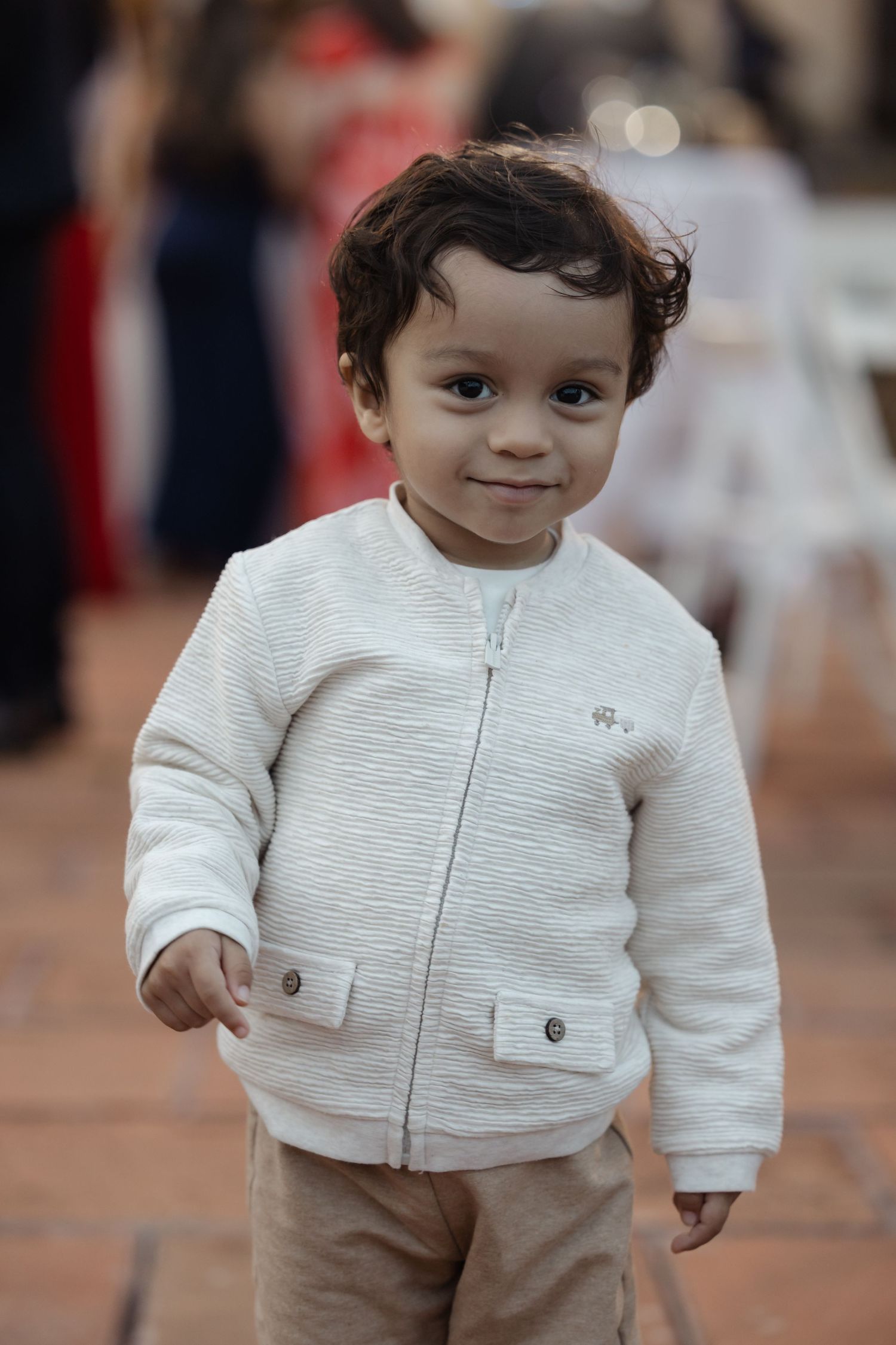 Adorable toddler in a white zip-up jacket smiles at the camera during a wedding reception.