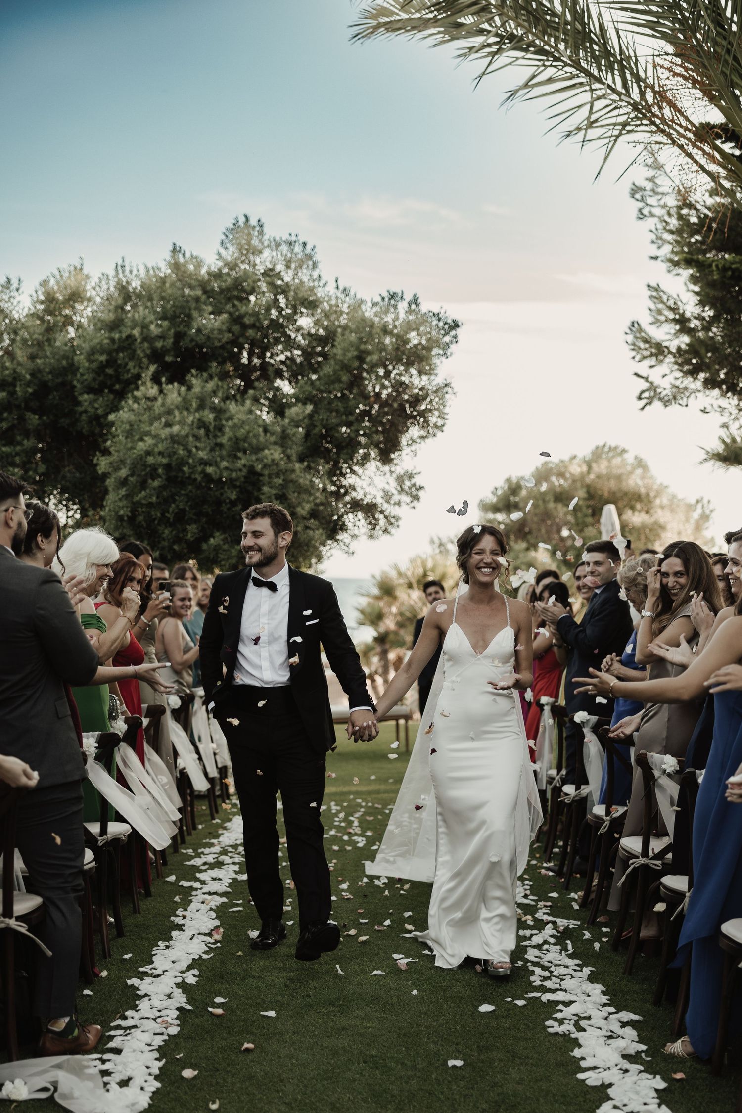 Happy newlyweds walk down the aisle as guests toss white rose petals at outdoor garden wedding.