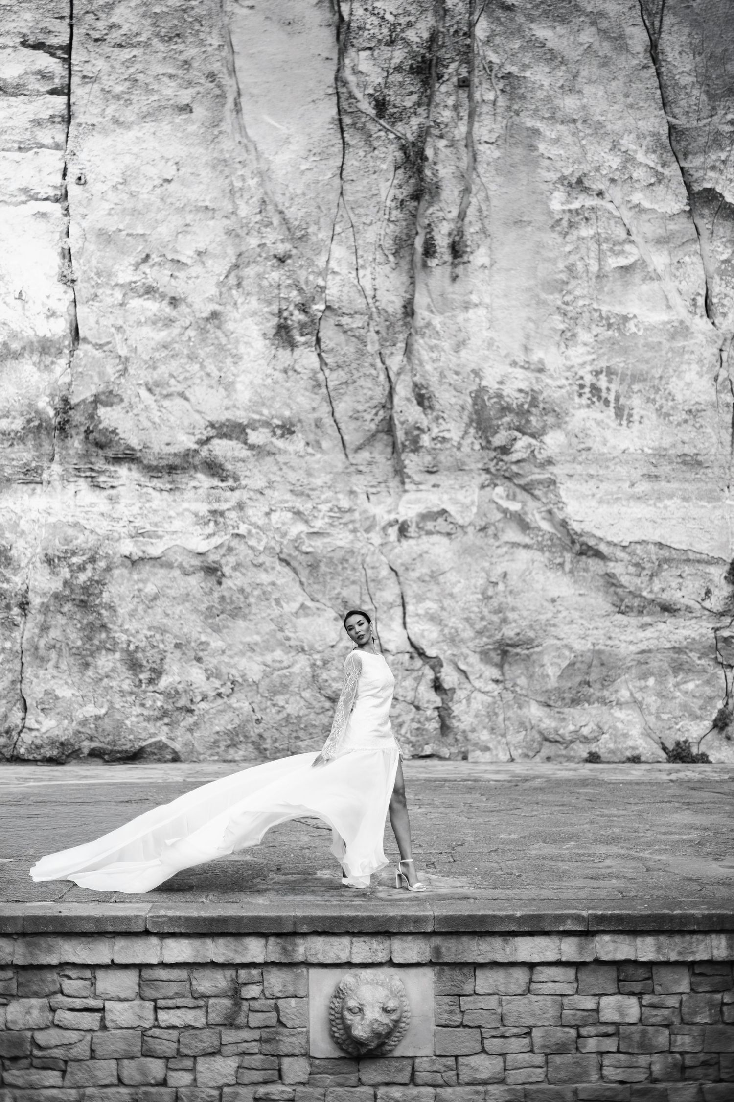 Bride in flowing white gown dancing dramatically before large stone wall in black and white.