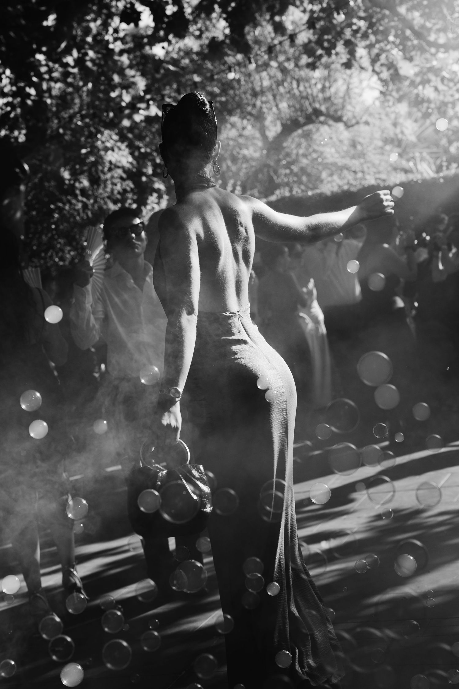 Black and white rear view of performer with bare back holding props surrounded by bokeh lights outdoors.