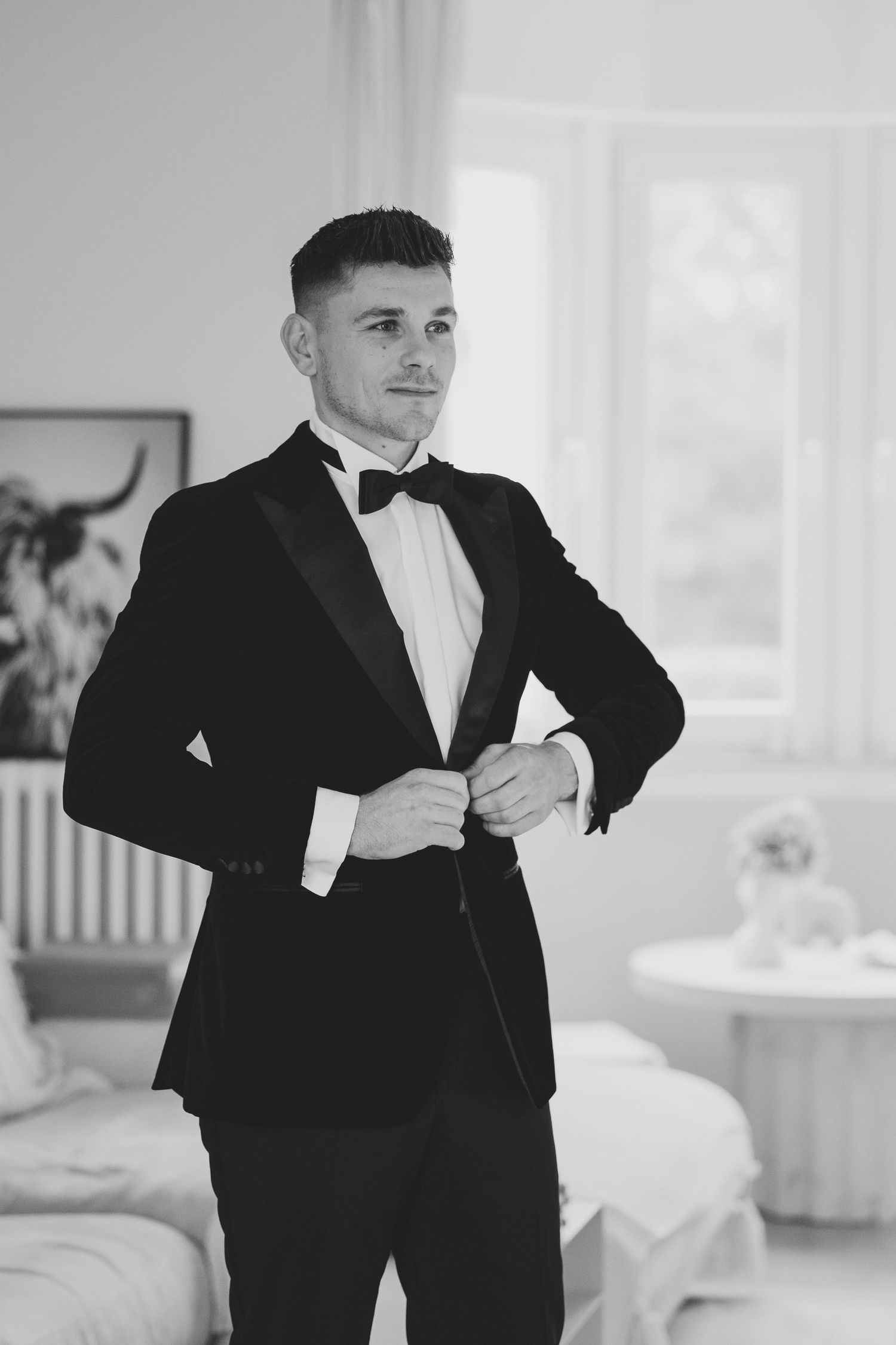 Dapper groom in black tuxedo with bow tie buttoning his jacket in a bright room, black and white photo.