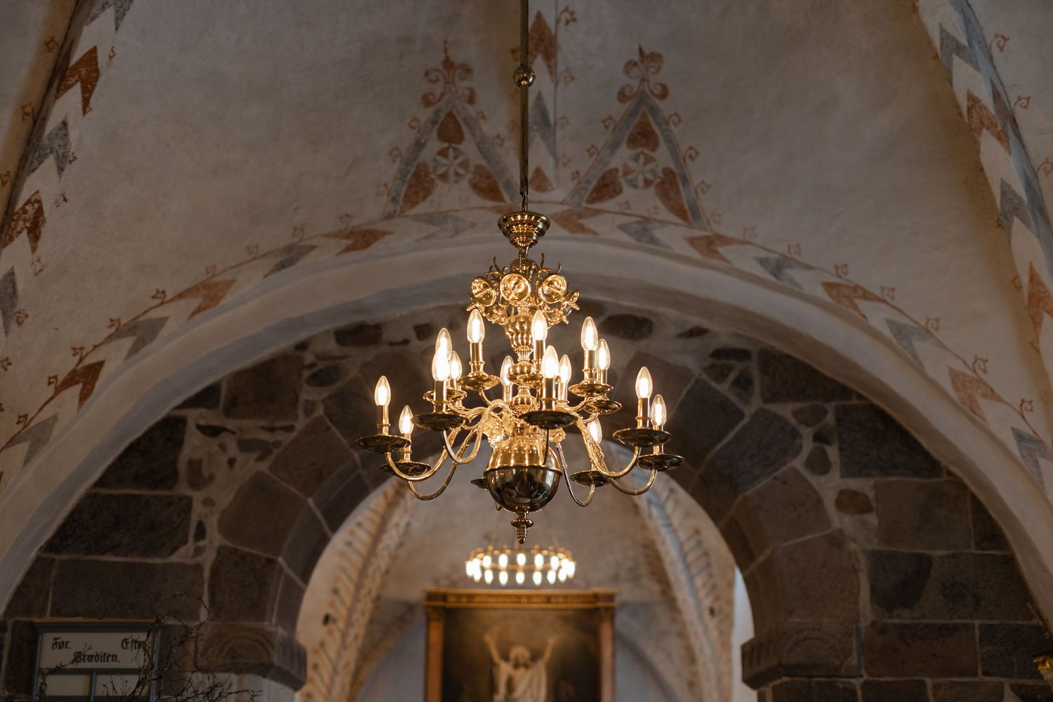 Gold chandelier with candle lights hanging under a stone arch with medieval frescoes in an old church.