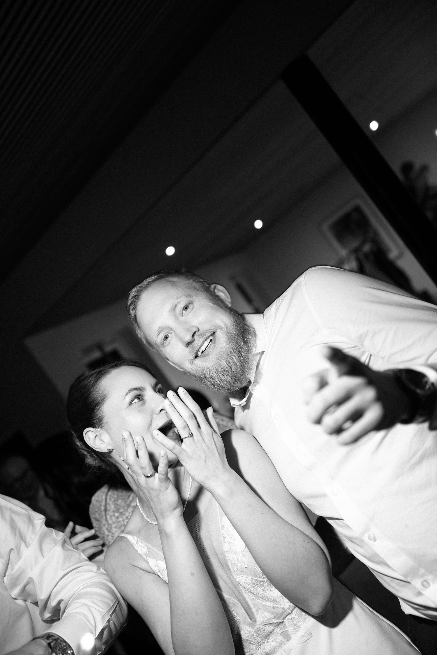 Excited bride covers her face with ringed hand while bearded groom gestures in black and white reception photo.
