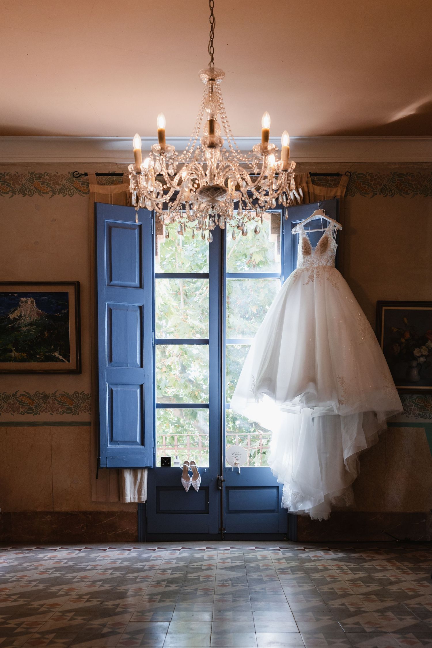 White wedding dress hanging from crystal chandelier near blue French doors in elegant vintage room.