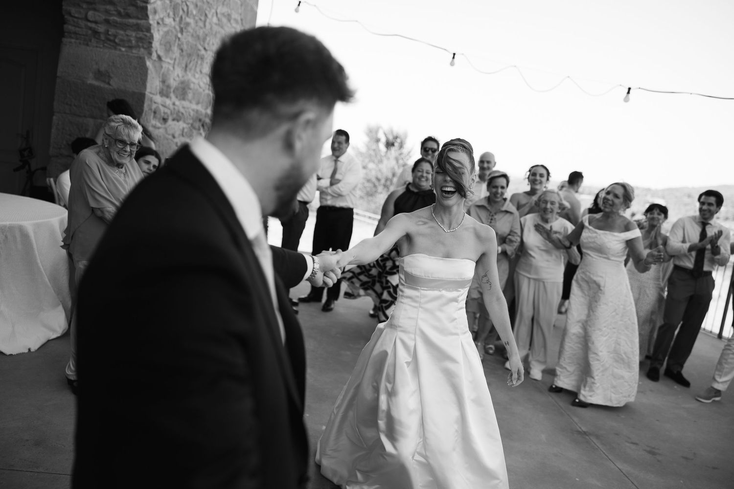 Black and white wedding photo of bride and groom dancing while guests clap and celebrate at outdoor reception.