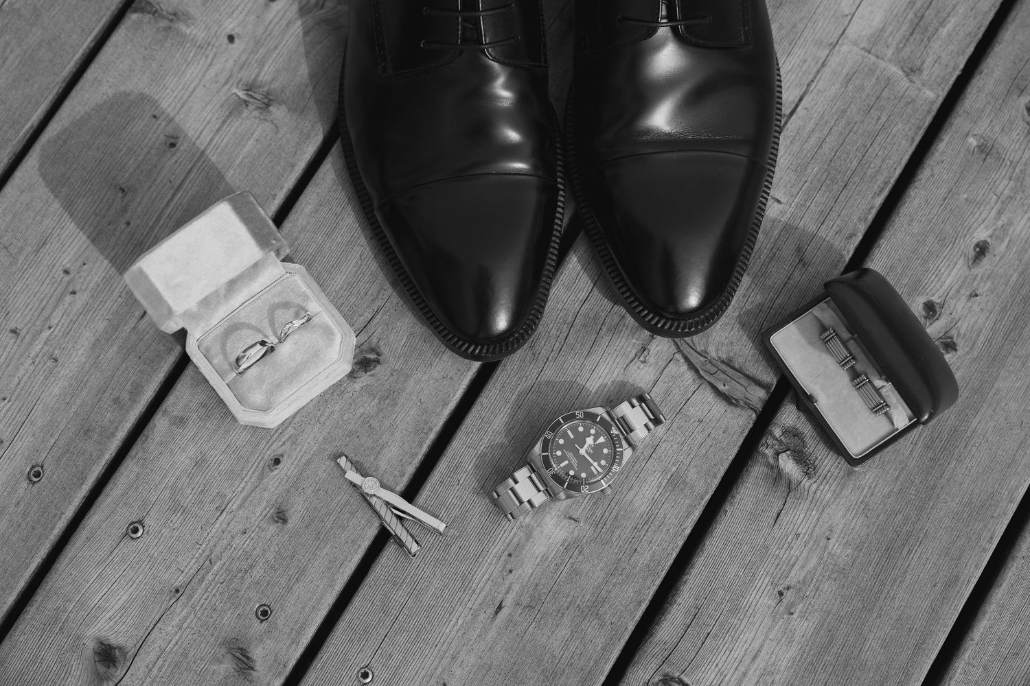 Black and white flat lay of mens dress shoes, cufflinks, watch, and ring boxes on wooden floor.