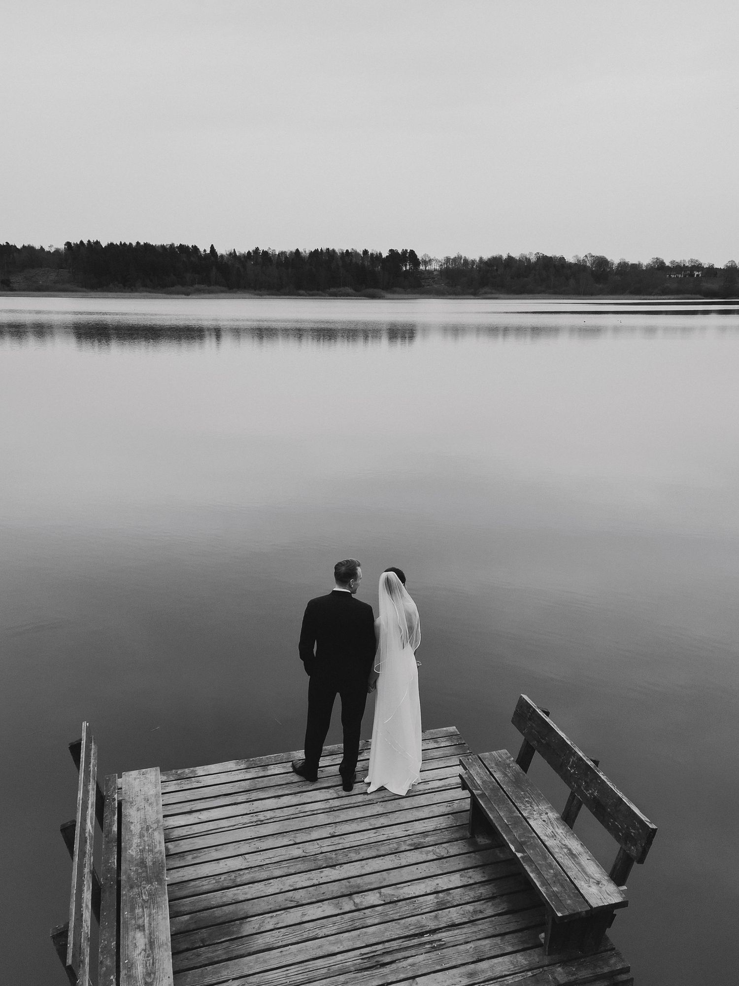 Black and white photo of bride and groom standing on a wooden dock overlooking a serene misty lake.