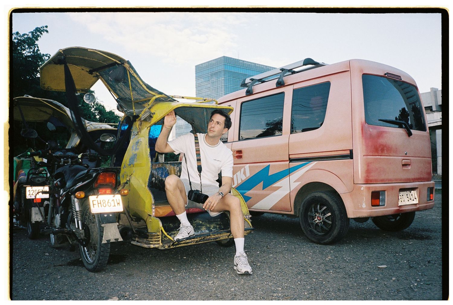 A young man sits casually on a colorful tuk-tuk beside a pink van in an urban parking area.