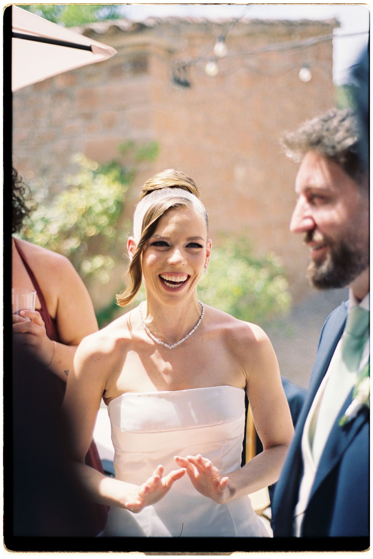 A joyful bride in a strapless white gown laughs with guests at an outdoor wedding reception.