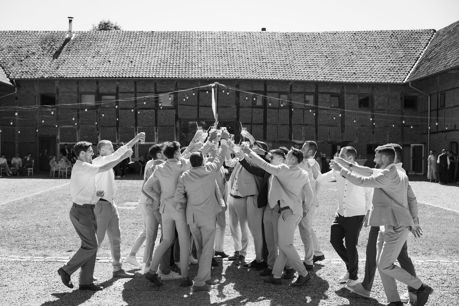 Groomsmen celebrating in a circle at a barn wedding venue, raising glasses in a joyful toast outdoors.