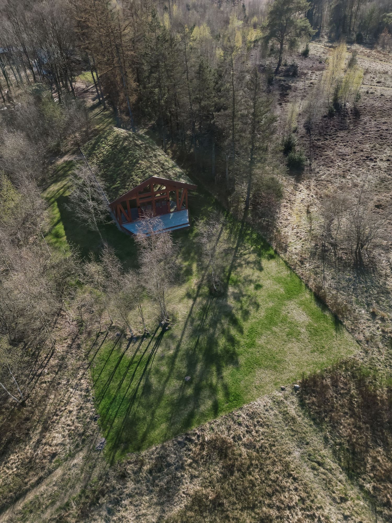 Aerial drone view of a rustic red cabin surrounded by forest trees with a green lawn clearing.
