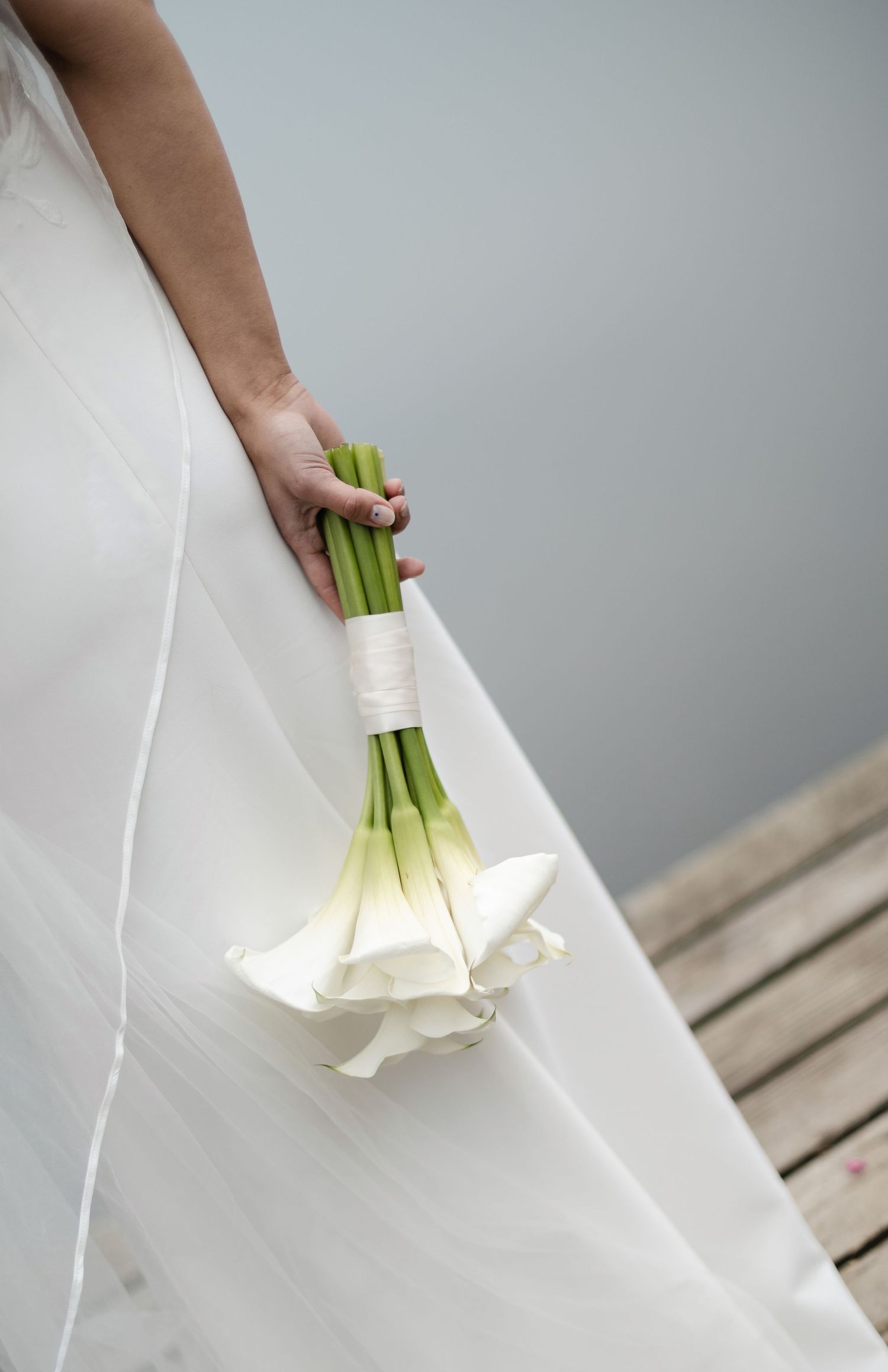 Bride in white wedding dress holding an elegant bouquet of white calla lilies tied with ribbon.