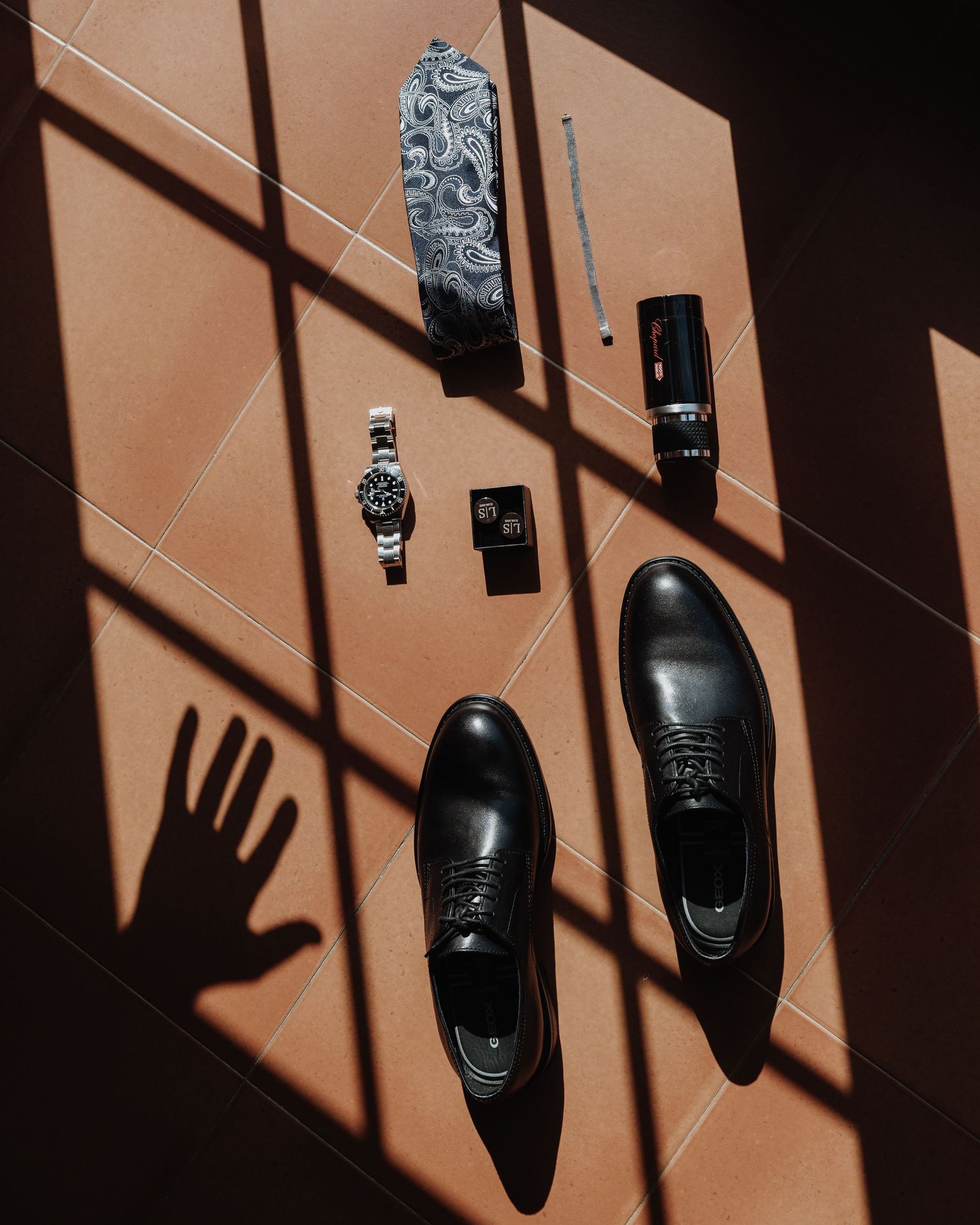 Flatlay of groom wedding accessories including black Oxford shoes, watch, tie, and cologne on tiled floor.