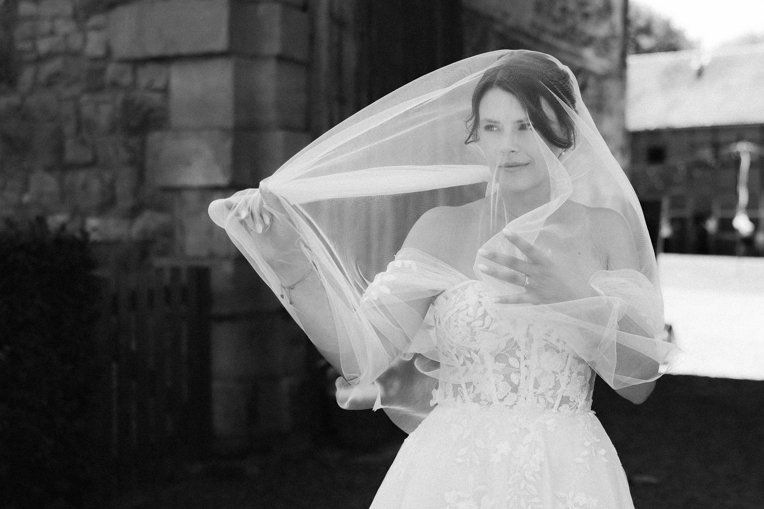 Black and white photo of a bride in a lace gown with veil blowing in the wind outside a stone building.