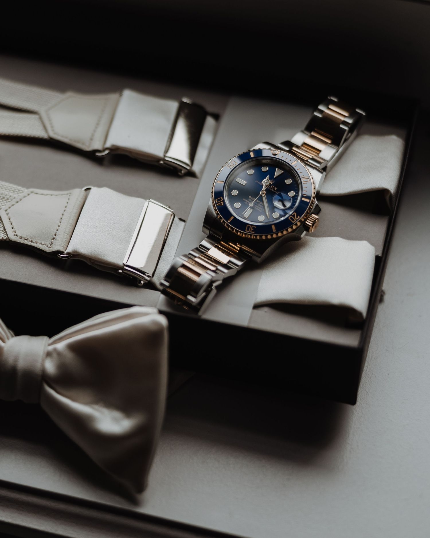 Luxury Rolex watch with blue dial displayed alongside bow tie and cufflinks on gray velvet tray.