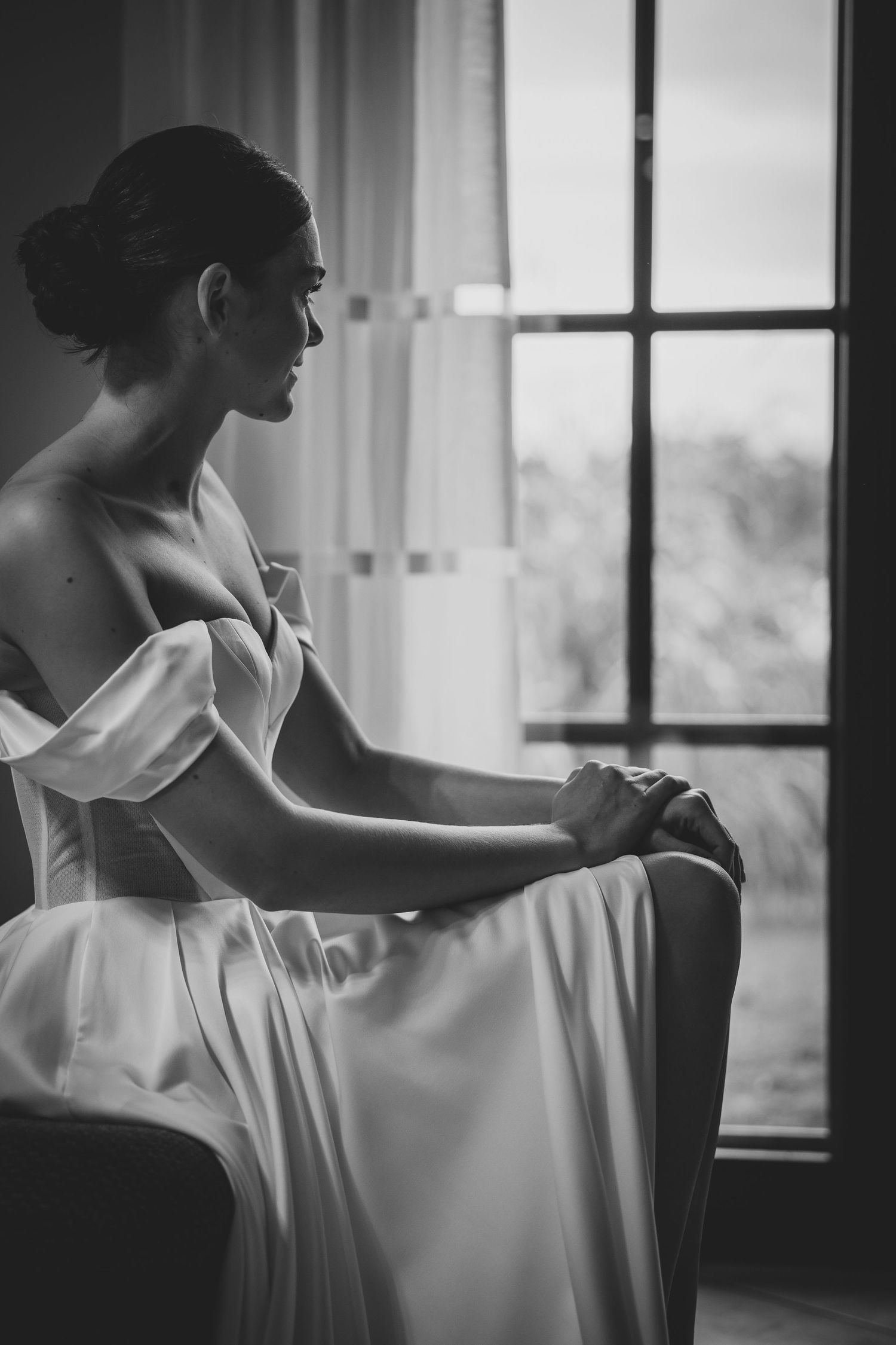 Bride in off-shoulder white gown sitting pensively by a large window, elegant black and white portrait.