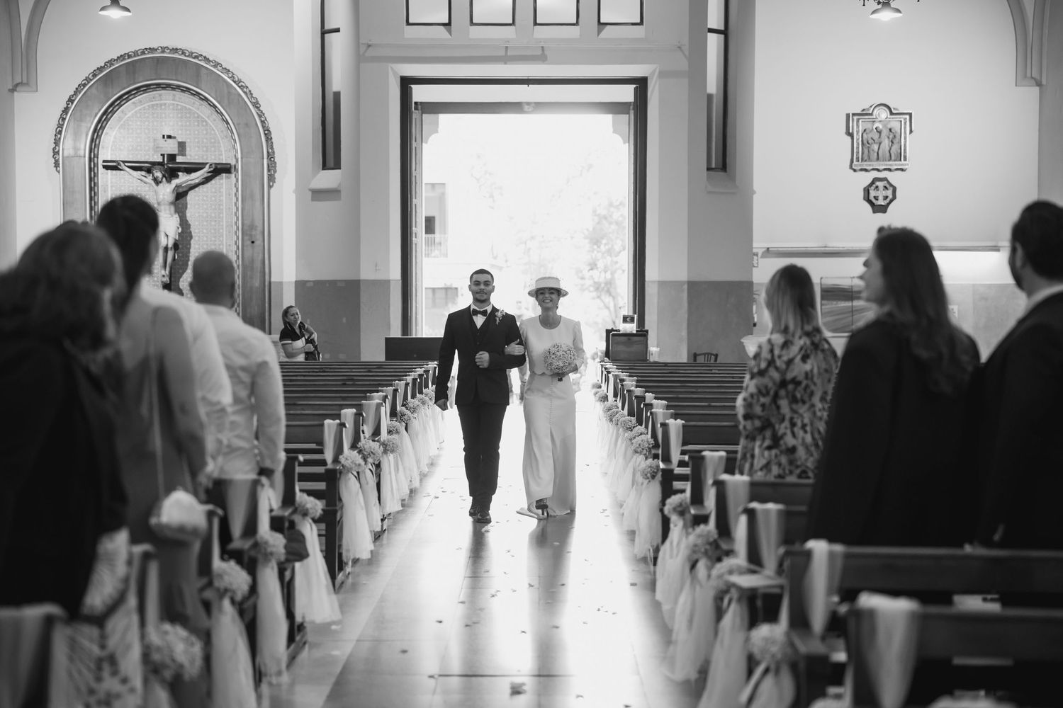 Black and white photo of a bride walking down the church aisle with escort during wedding ceremony.