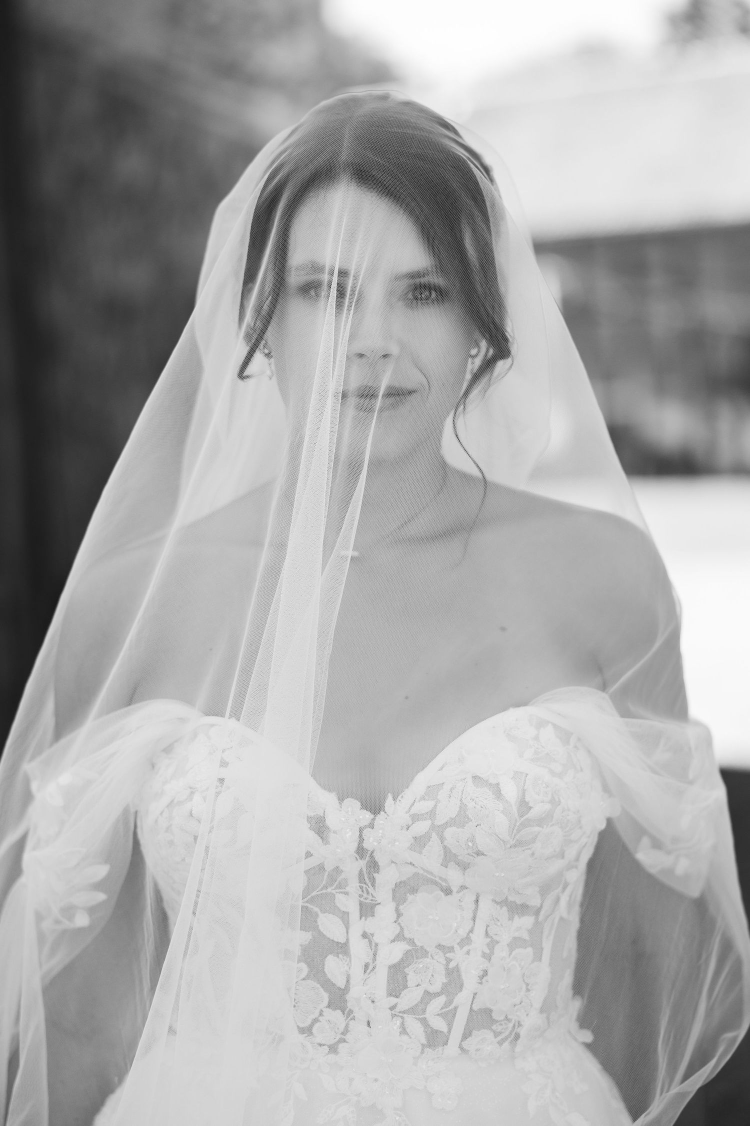 Elegant bride in lace off-shoulder gown with flowing veil in stunning black and white portrait.
