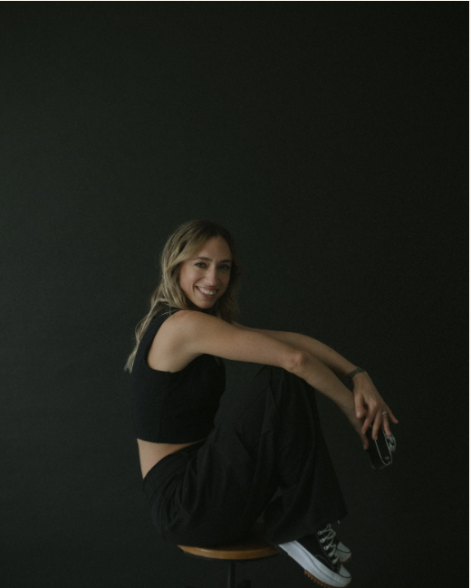 A smiling young woman in black athletic wear sits relaxed against a dark studio background.