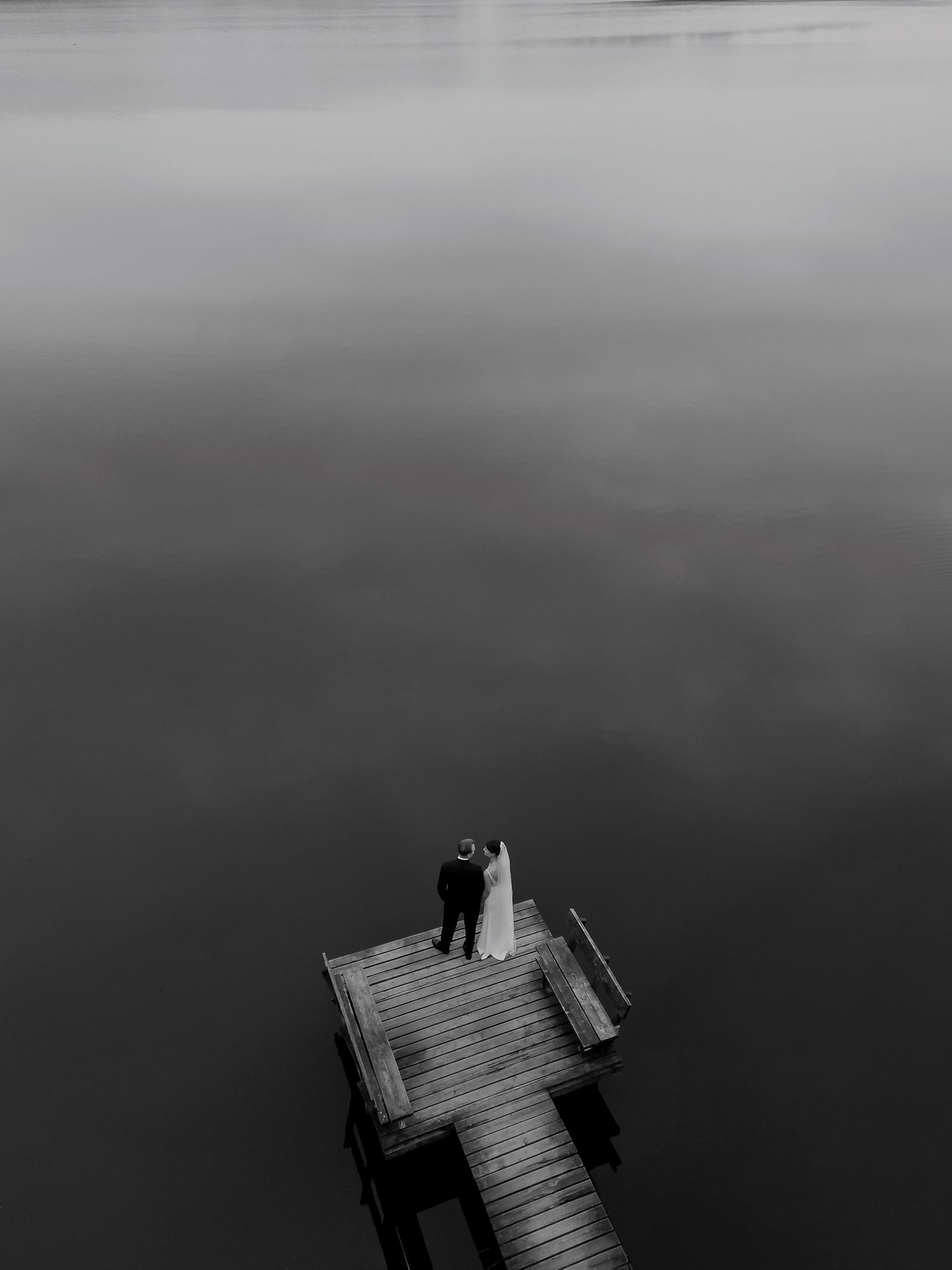 A couple stands on a wooden dock over calm water in a stunning black and white aerial photo.