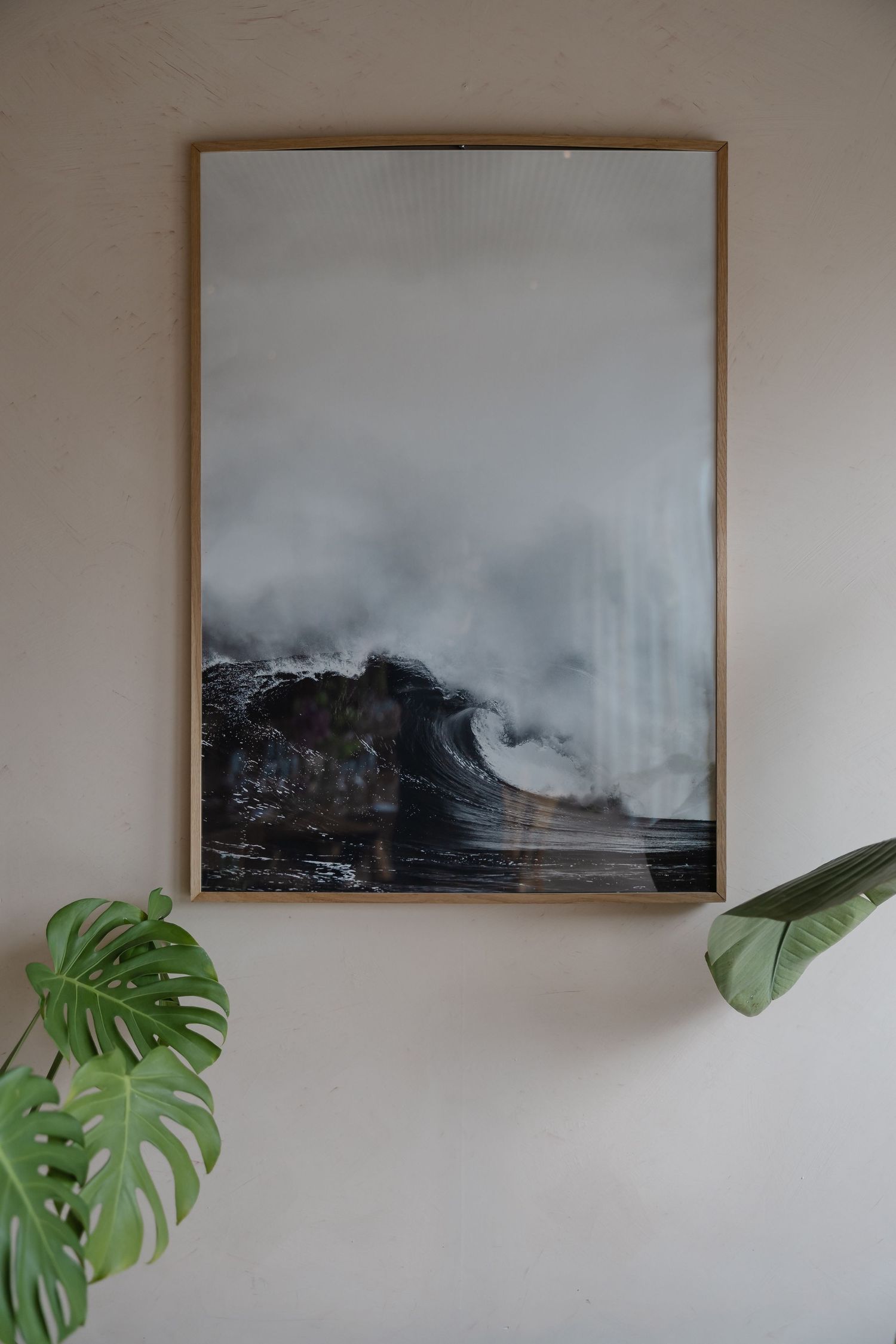 Framed ocean wave art print in gold frame on beige wall with monstera leaves.
