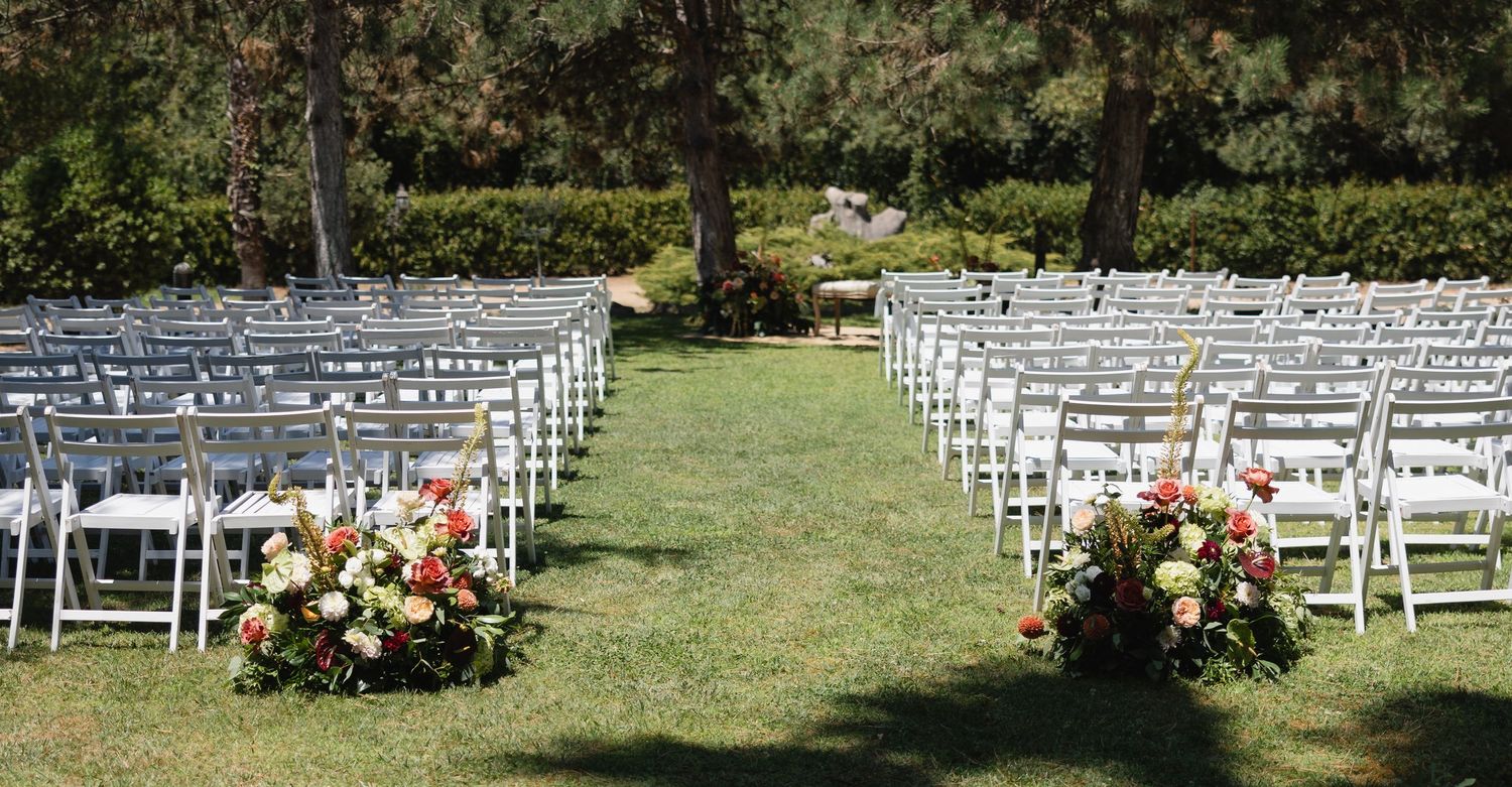 Outdoor wedding ceremony setup with white chairs and colorful floral arrangements on green lawn.