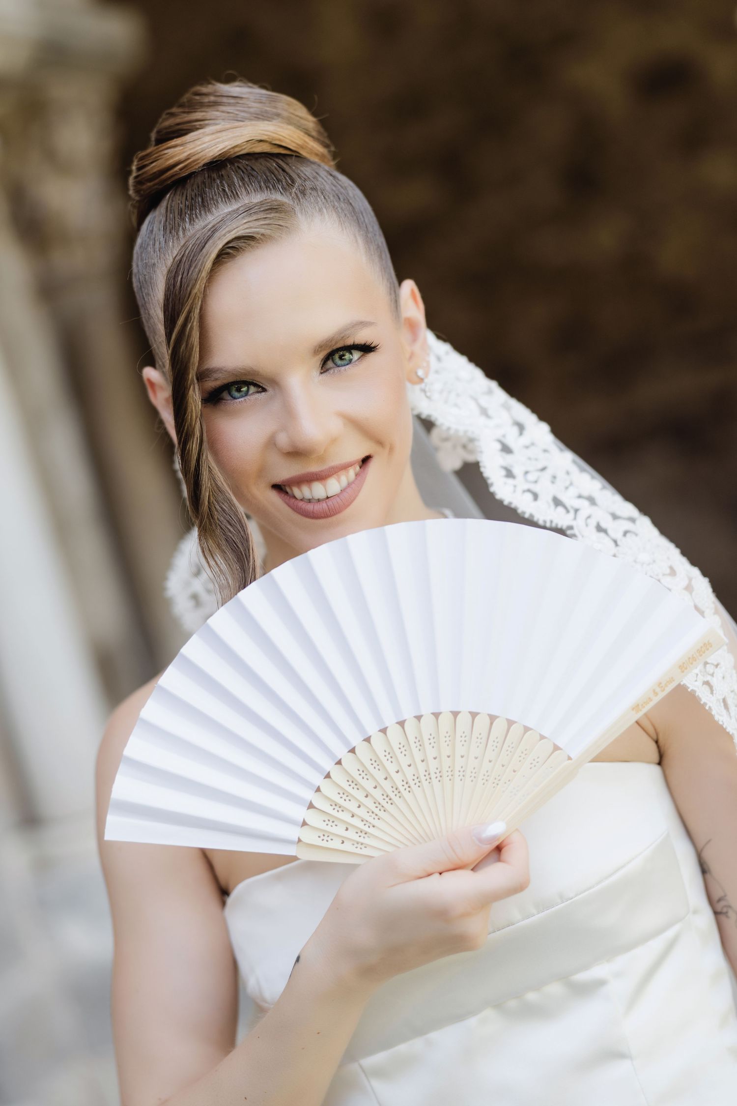 Smiling bride holding white hand fan wearing elegant updo hairstyle and lace veil at outdoor wedding venue.