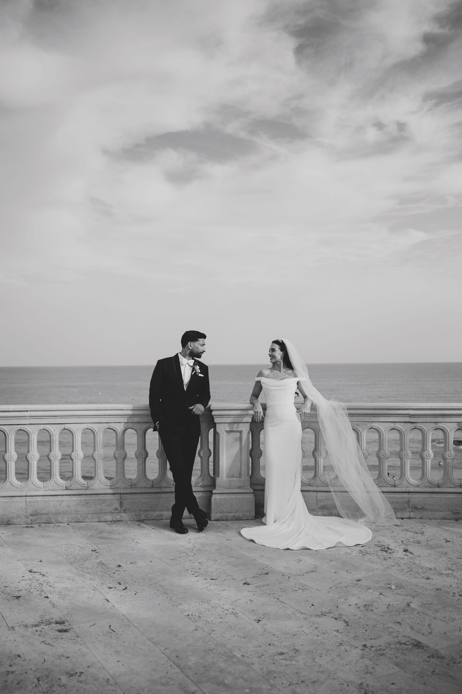Black and white photo of bride and groom standing on oceanfront terrace balustrade under cloudy sky.