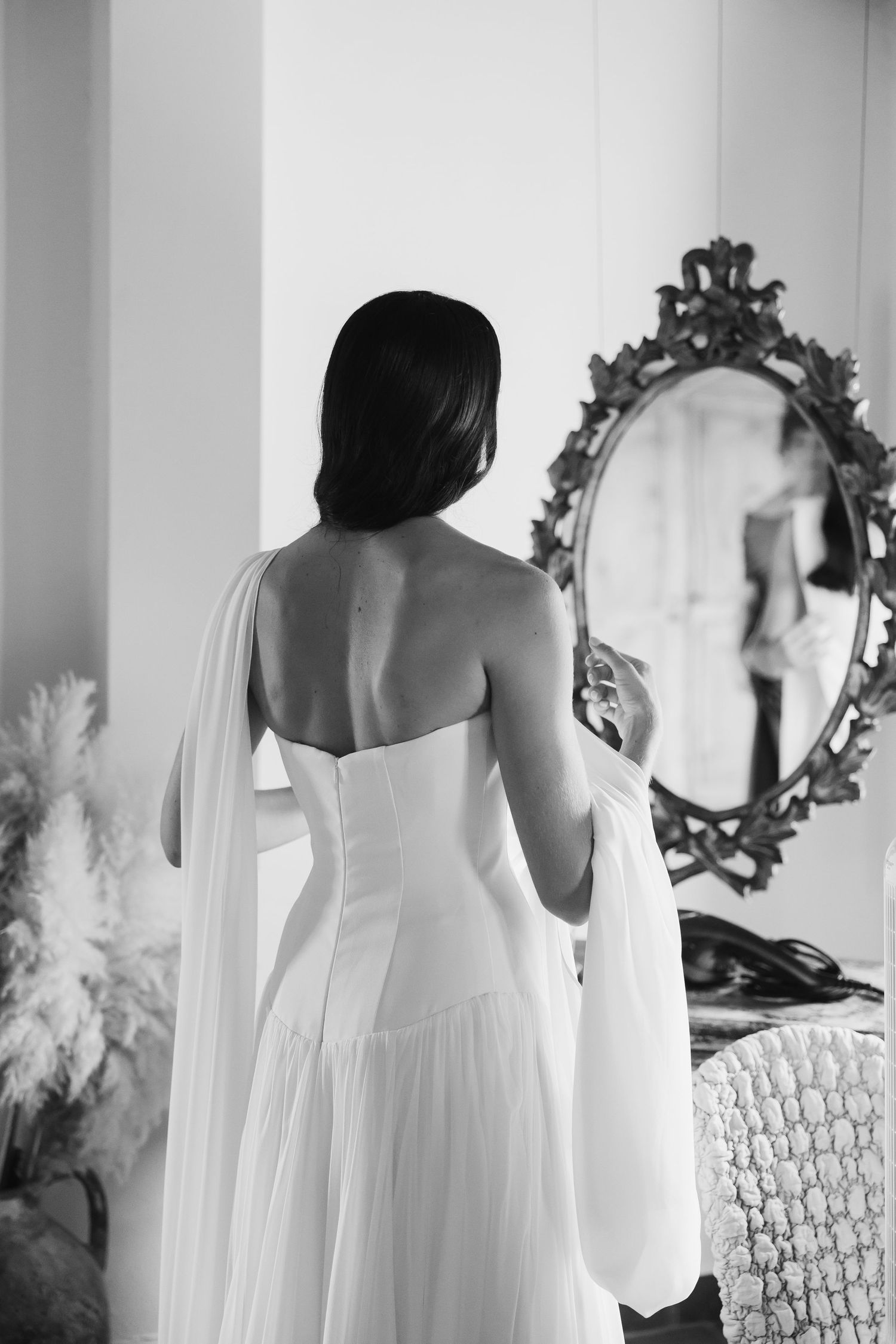 Black and white photo of a bride in a strapless wedding gown viewed from behind, facing an ornate mirror.