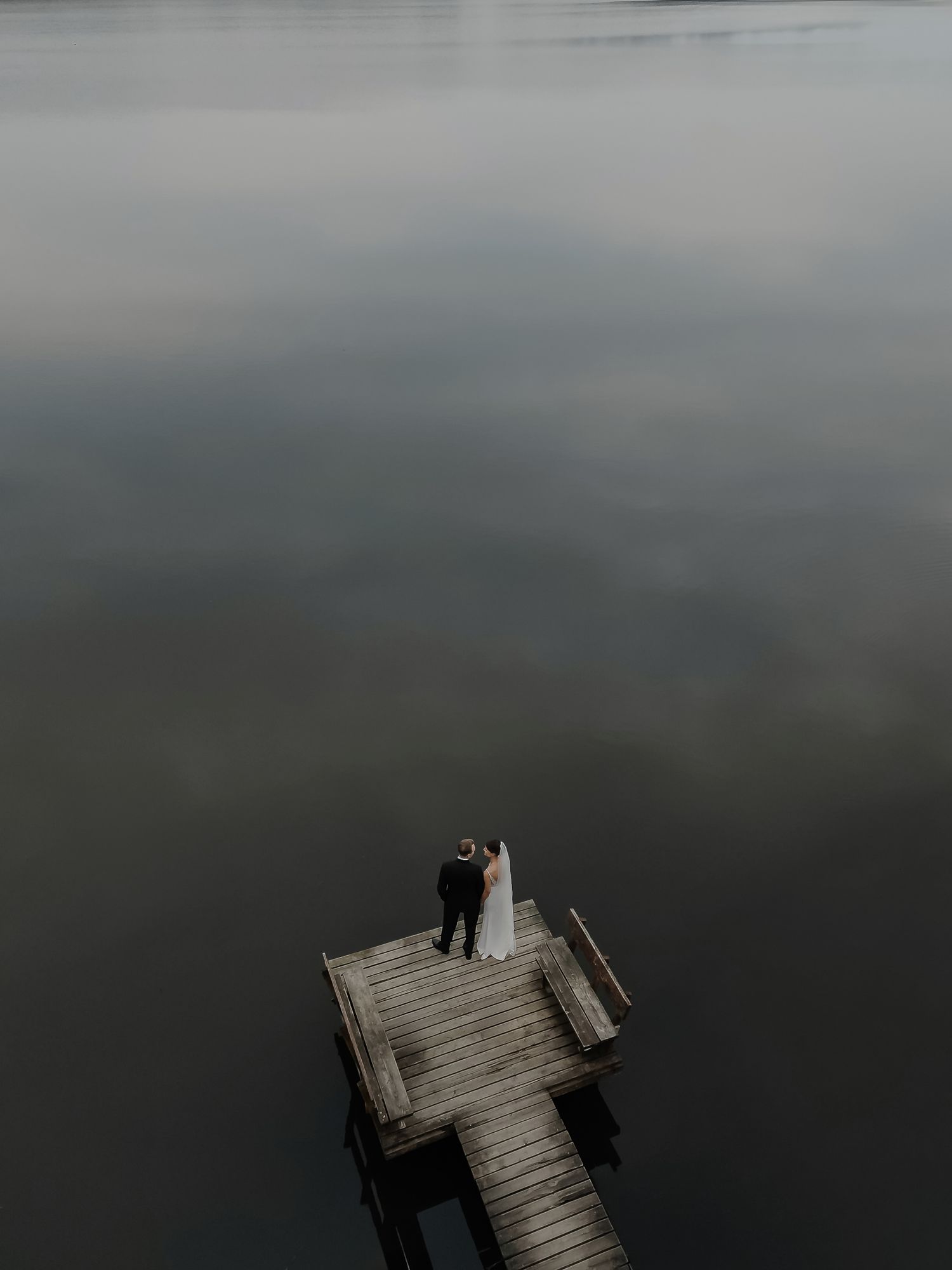 Aerial view of bride and groom standing on a wooden dock surrounded by still water.