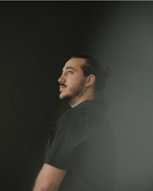 A young man with a bun hairstyle wearing a black t-shirt looks upward in a dramatic dark studio portrait.