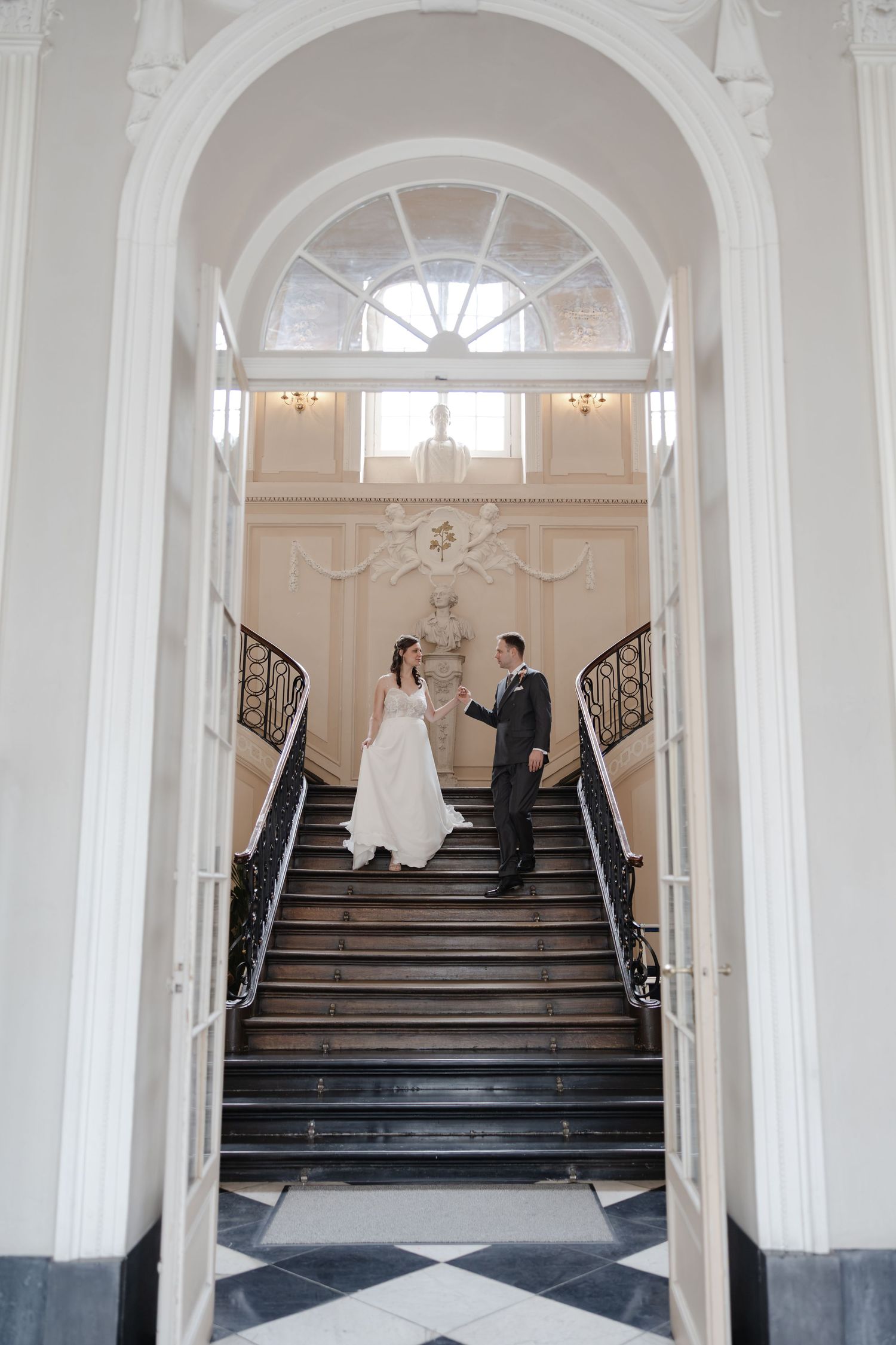 Bride and groom hold hands on grand staircase in elegant white venue with arched doorway and chandelier.