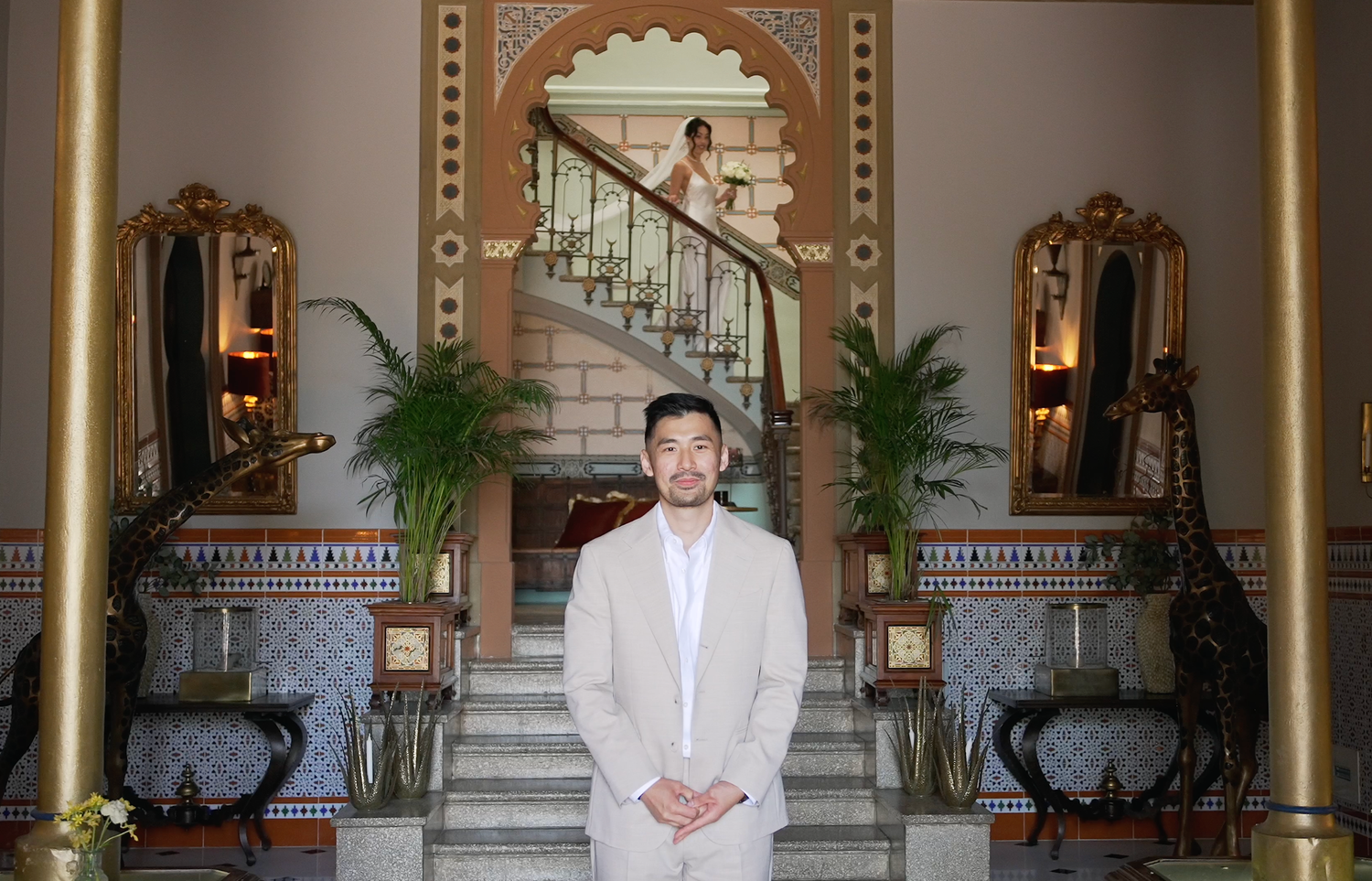 A man in a beige suit stands in an elegant ornate hotel lobby with grand staircase.