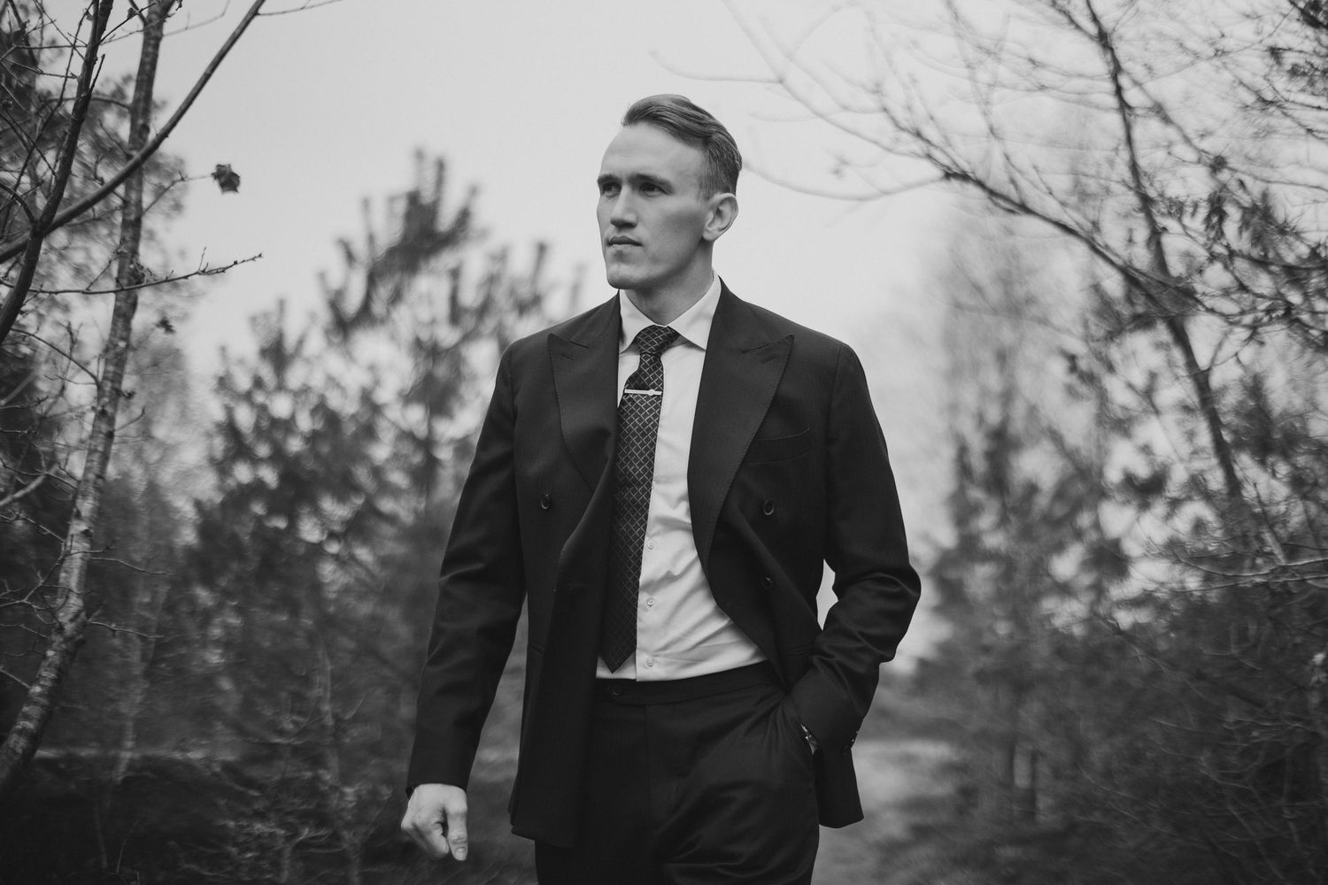 Elegant groom in black suit standing outdoors among trees in a black and white wedding portrait.